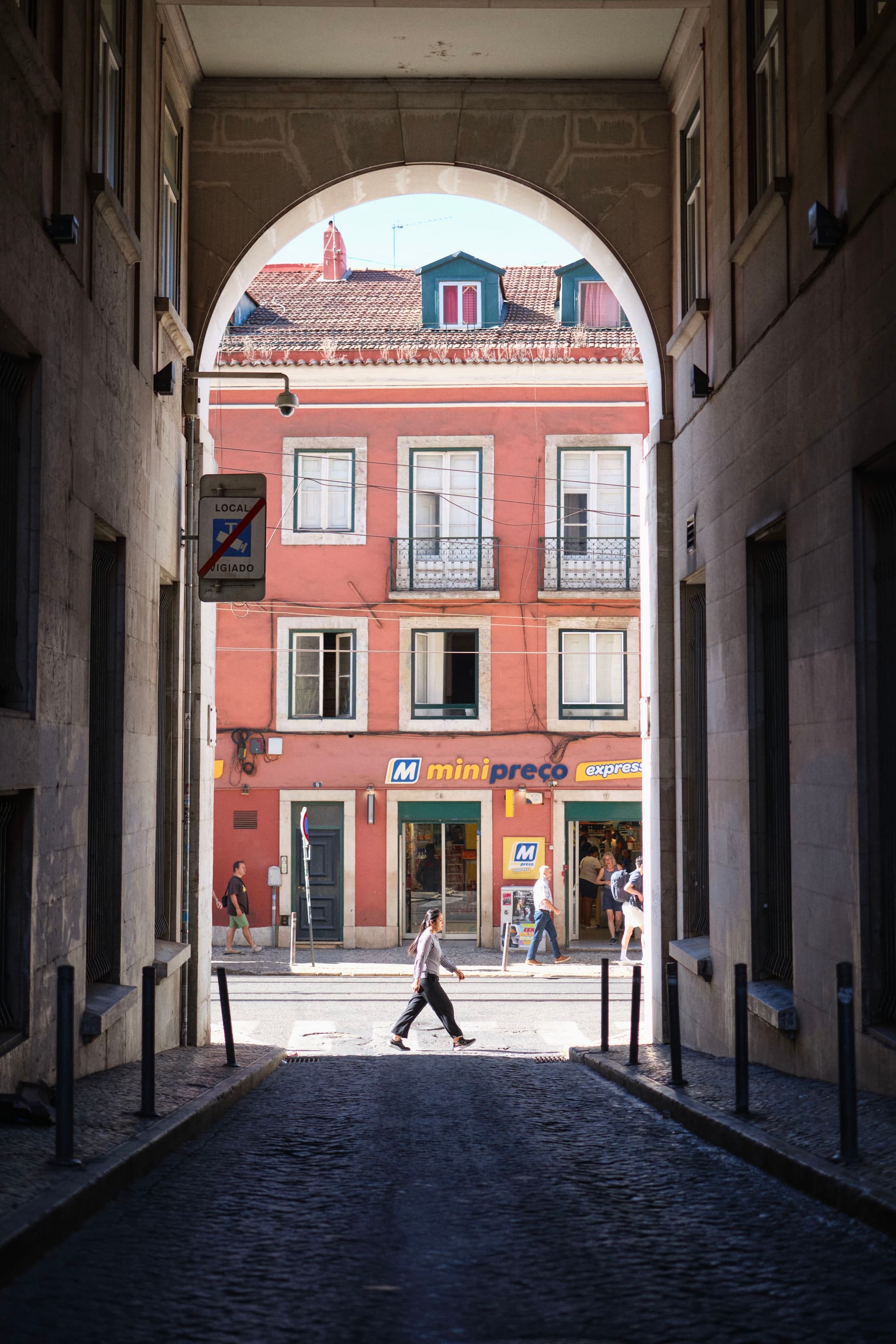 A person walks across a sunlit street, framed by a shadowy archway, with colorful buildings in the background