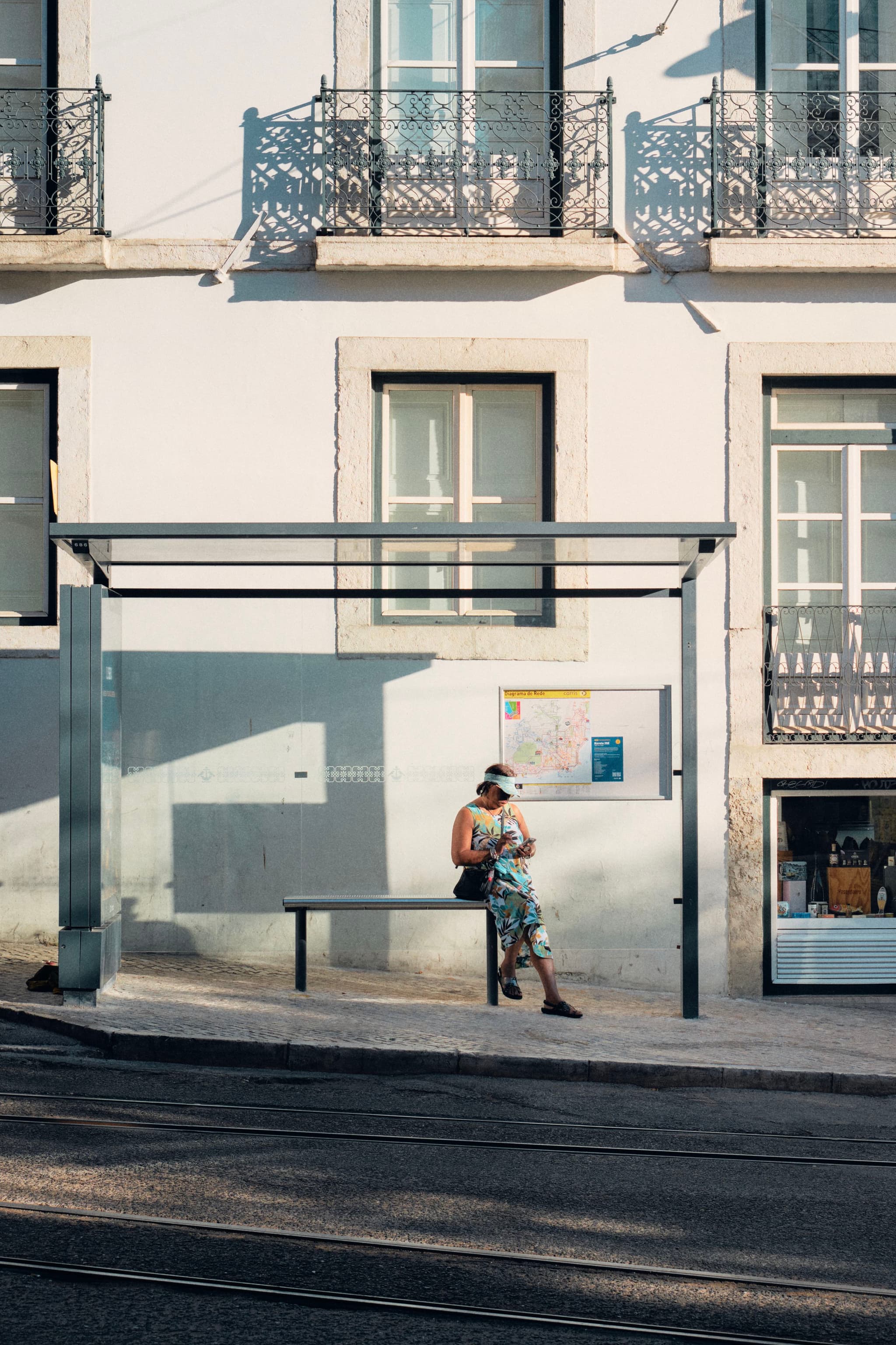 A person sits at a bus stop in front of a building with large windows and ornate railings, bathed in sunlight