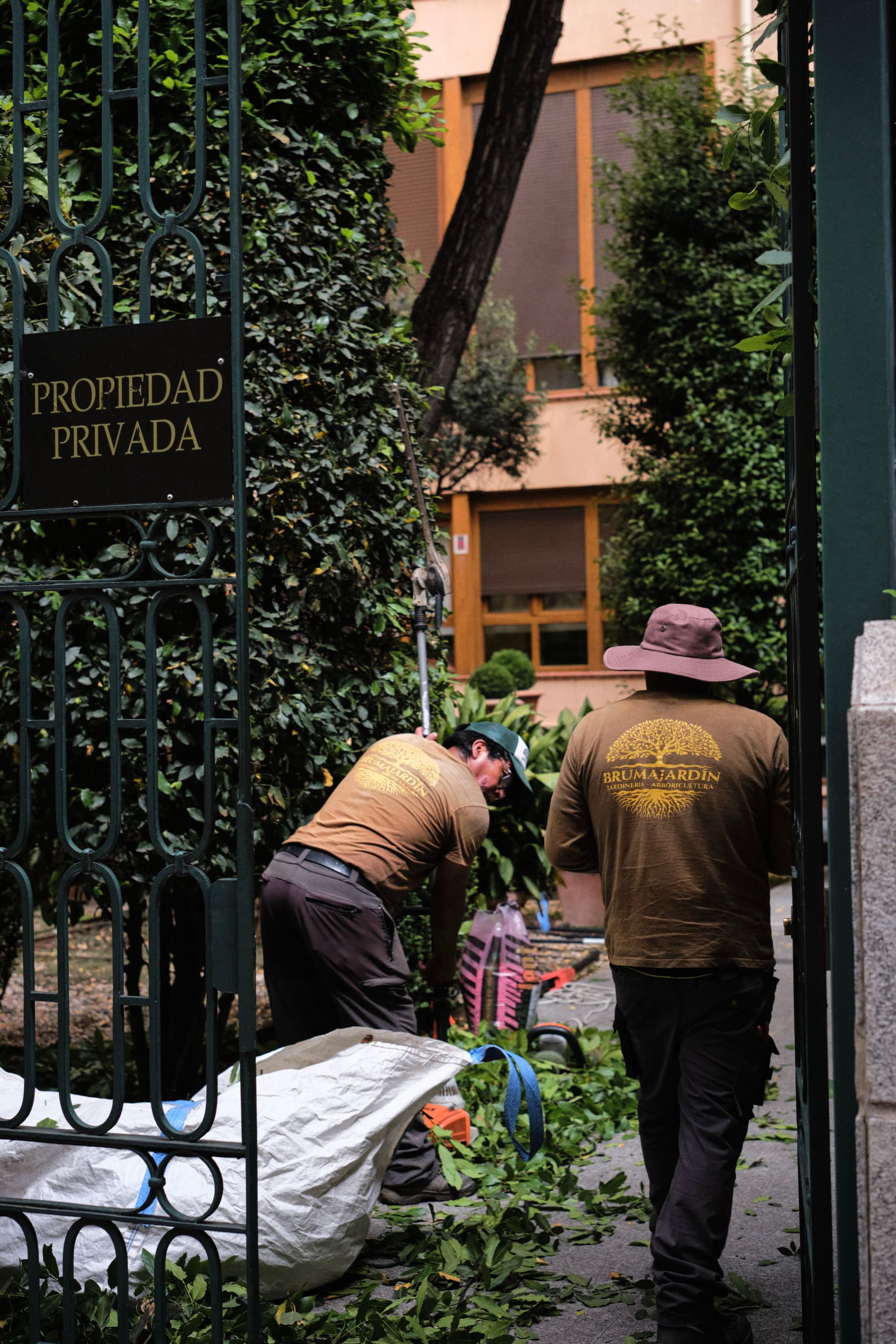 Two people working in a garden, surrounded by greenery, with a sign on a gate that reads PROPIEDAD PRIVADA
