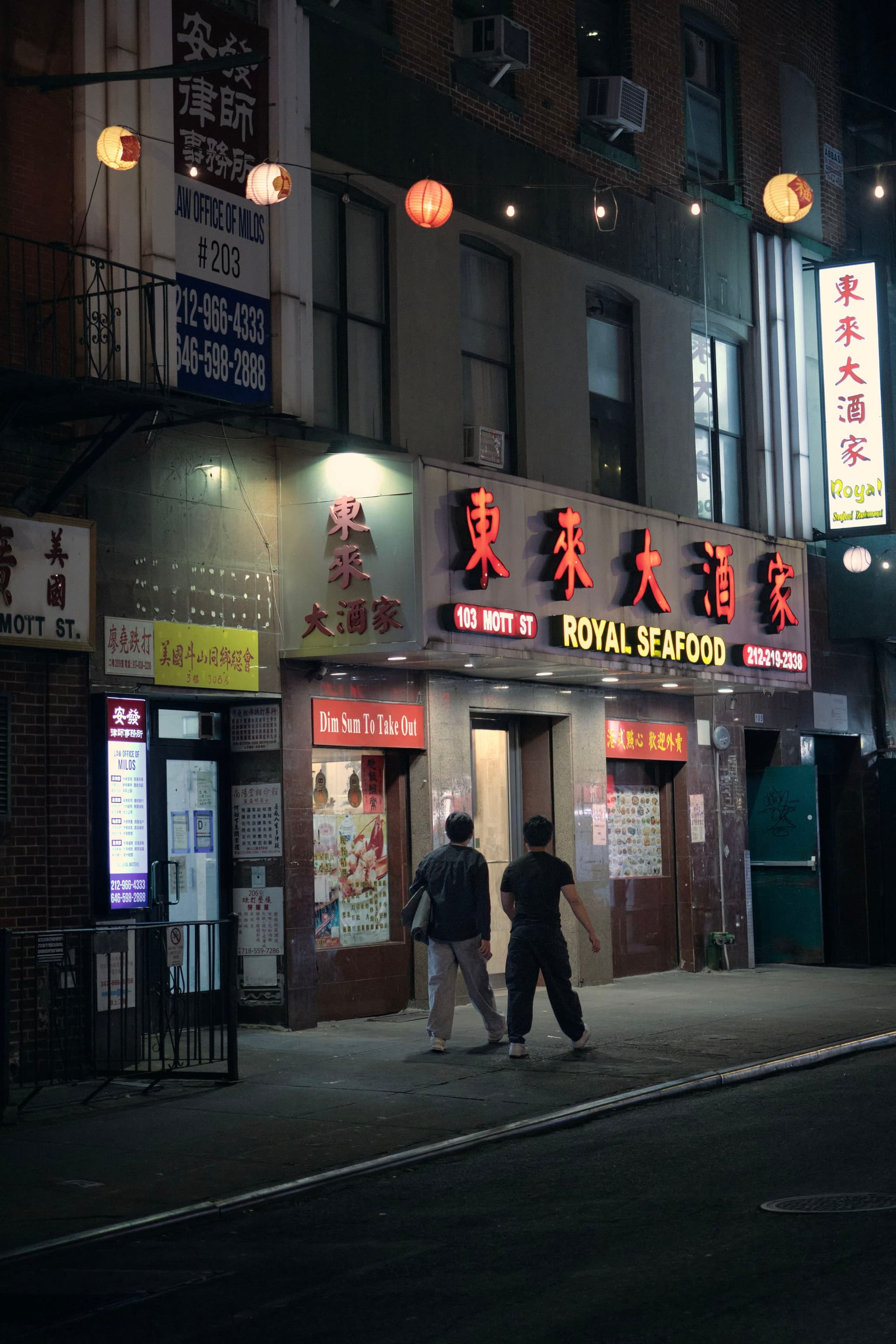 A dimly lit street scene featuring a restaurant with illuminated signs in Chinese characters and Royal Seafood in English. Two people are walking on the sidewalk in front of the establishment