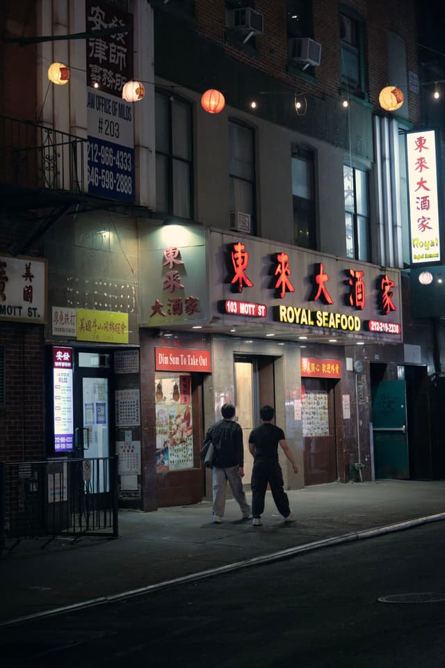 A dimly lit street scene featuring a restaurant with illuminated signs in Chinese characters and Royal Seafood in English. Two people are walking on the sidewalk in front of the establishment