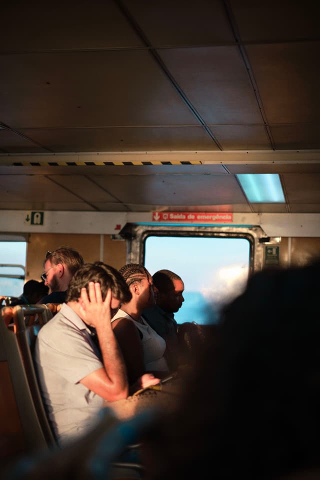 People sitting inside a dimly lit vehicle, possibly a train or bus, with sunlight streaming through a window