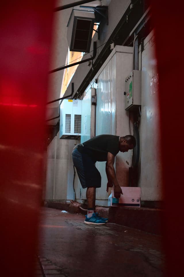 A person is bending down in a narrow alleyway, surrounded by walls and air conditioning units, with a red object partially framing the scene