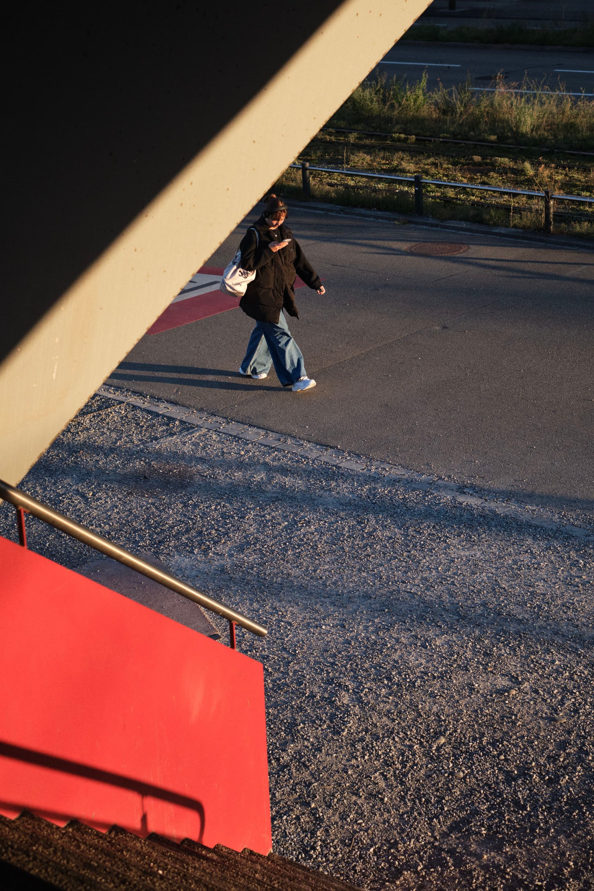 A person walking on a paved path, viewed from above, with a red structure and shadows in the foreground