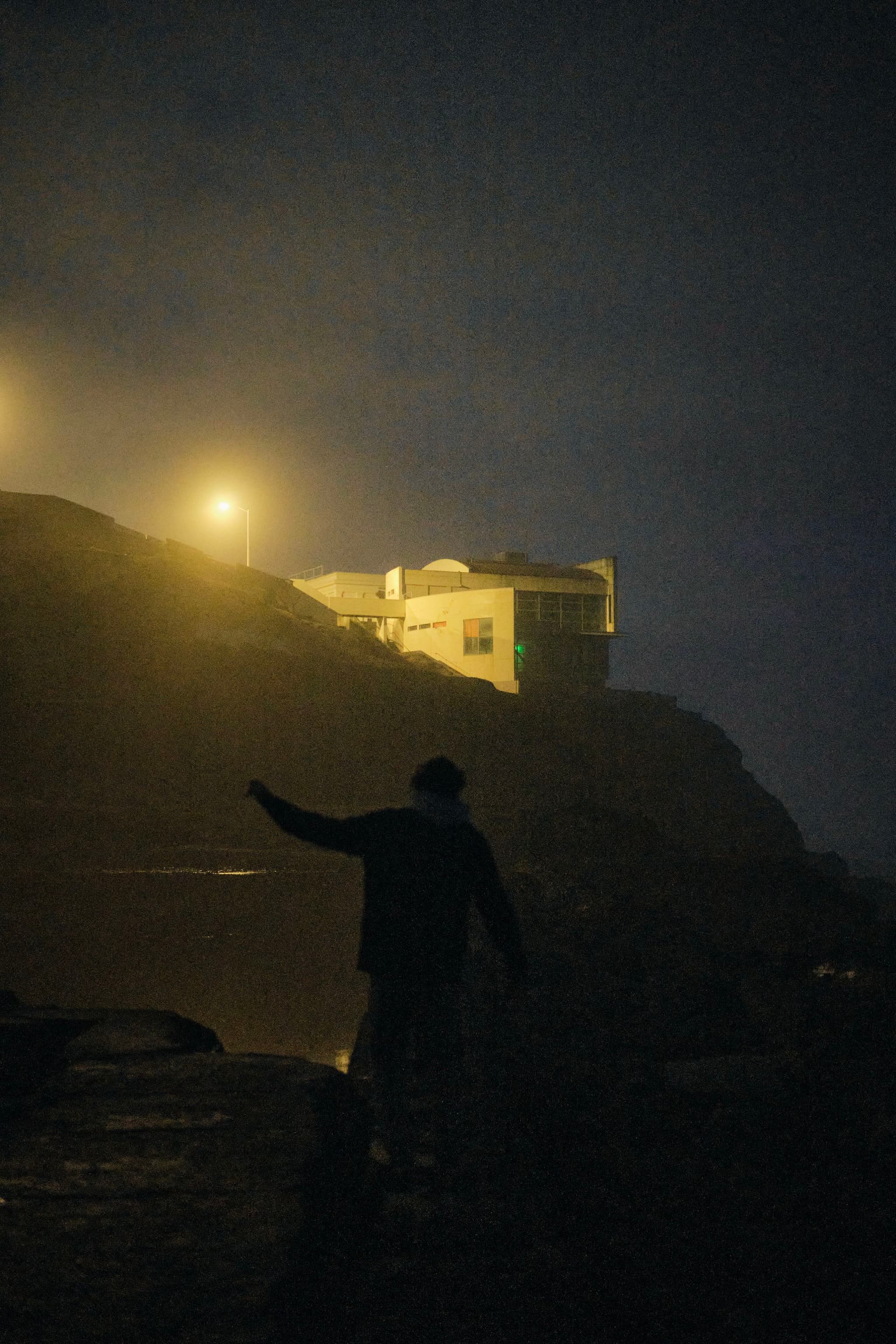 A silhouette of a person walking near a rocky area at night, with a building illuminated by streetlights in the background