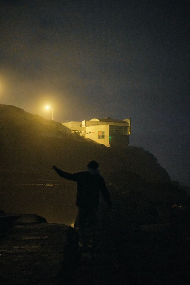 A silhouette of a person walking near a rocky area at night, with a building illuminated by streetlights in the background