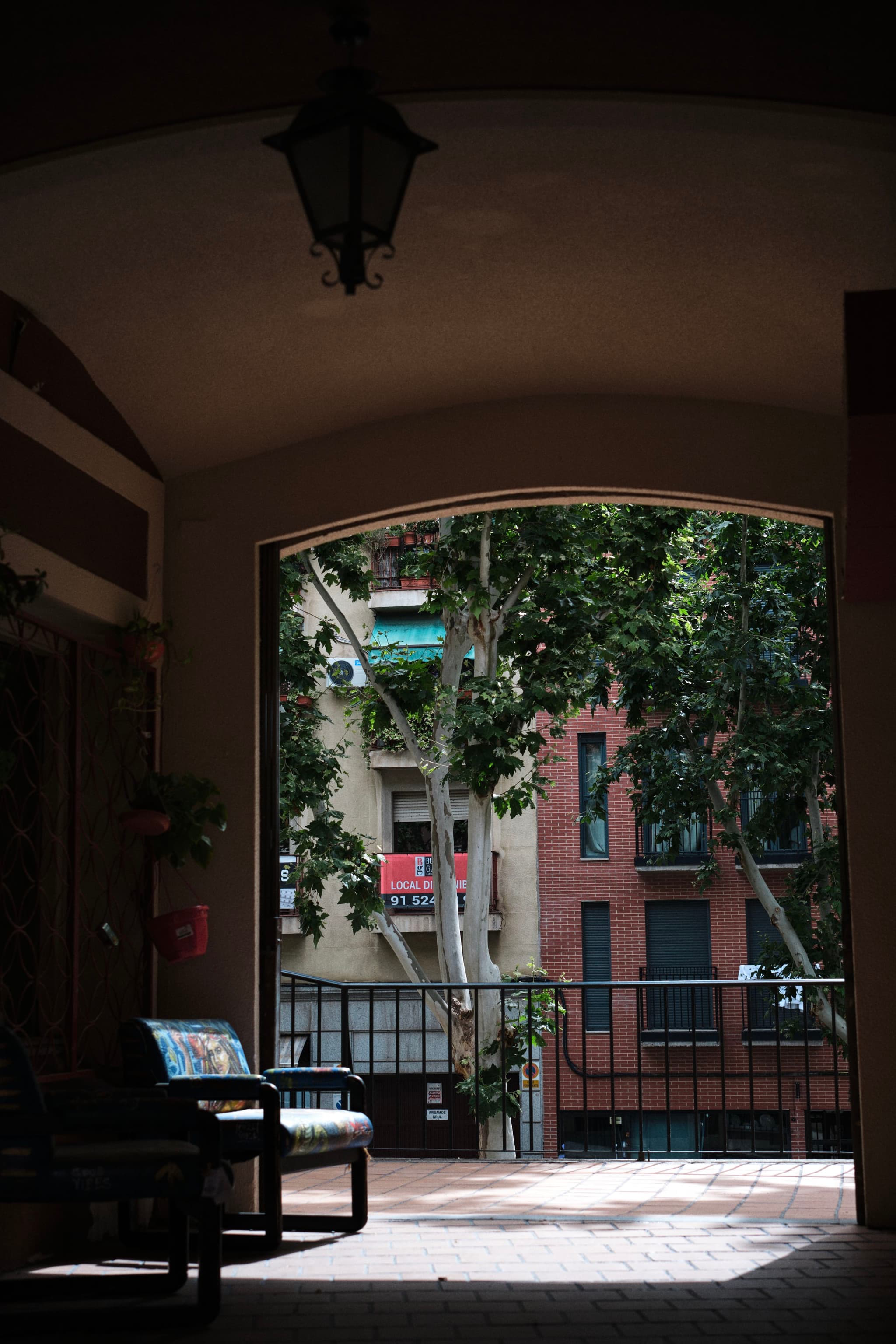 A shaded archway leads to an outdoor area with a bench, overlooking a view of trees and brick buildings