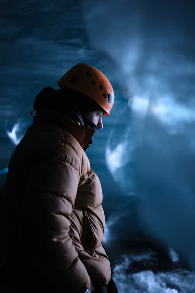 A person wearing a helmet and winter jacket stands in an icy cave, illuminated by blue light