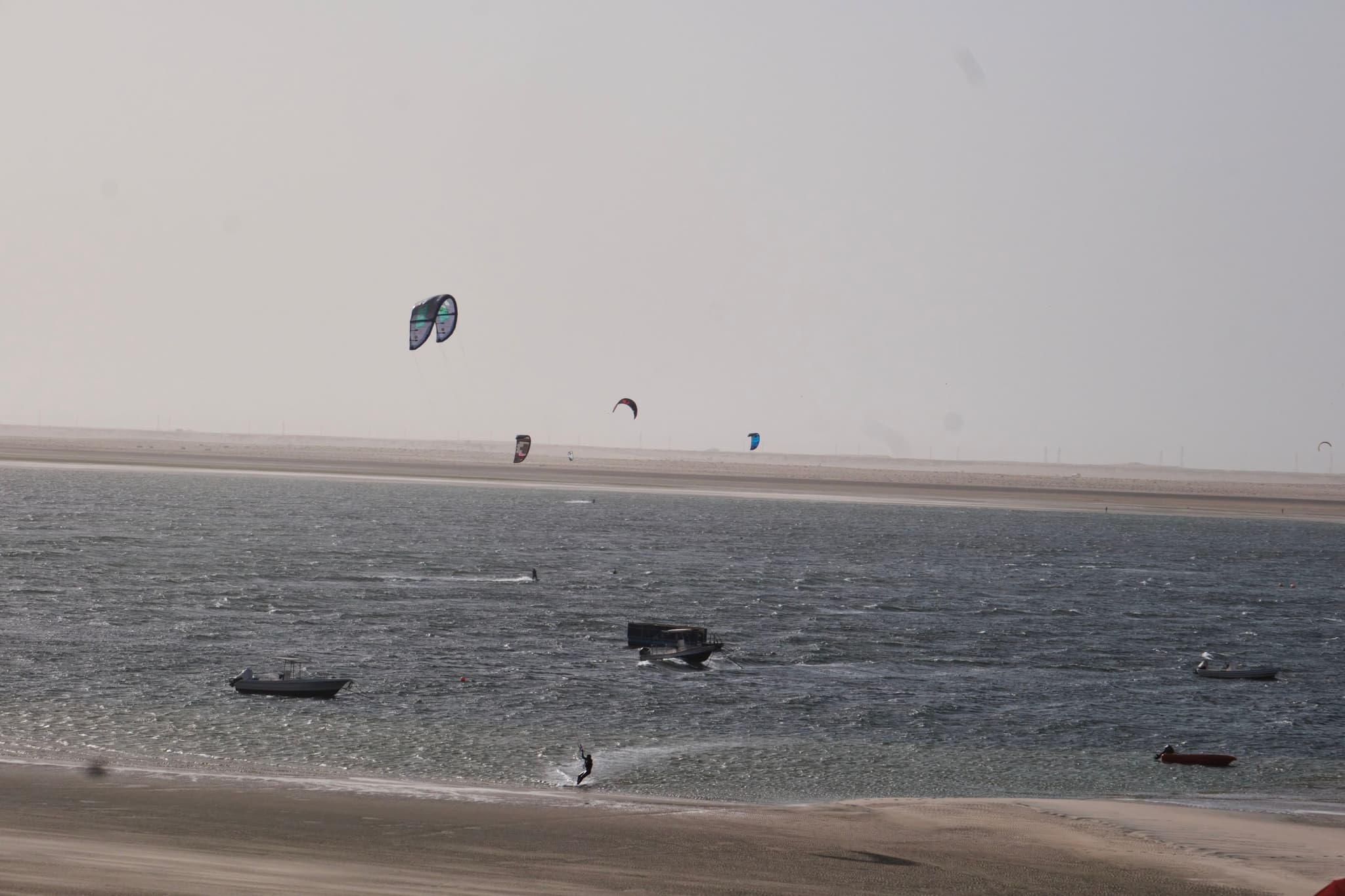 A beach scene with several people kite surfing on the water, small boats nearby, and a sandy shoreline