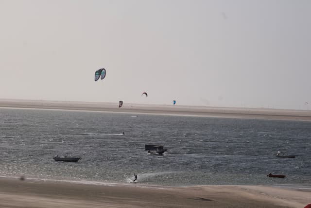 A beach scene with several people kite surfing on the water, small boats nearby, and a sandy shoreline