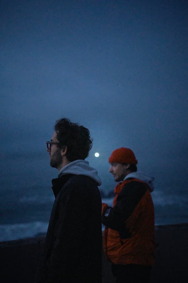 Two people stand near a body of water at dusk, with one wearing glasses and a hoodie, and the other in an orange jacket and beanie. The moon is visible in the cloudy sky