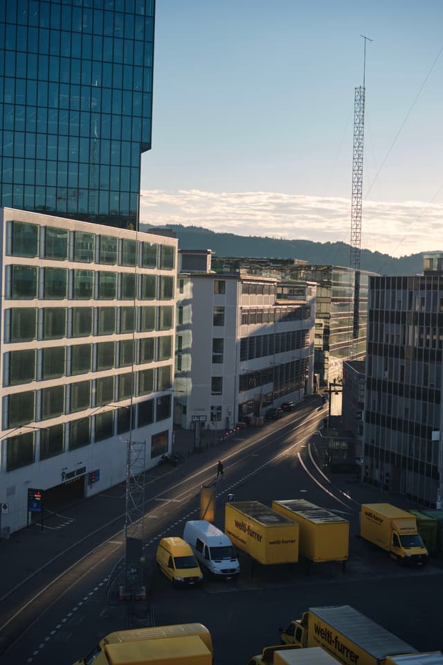 A city street scene with modern buildings, several yellow delivery trucks, and a clear sky in the background