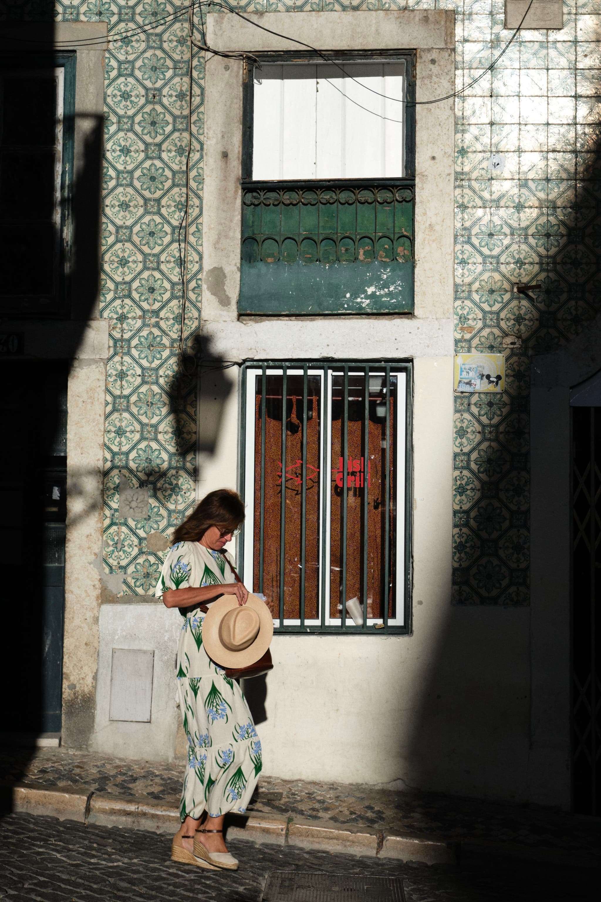 A person in a patterned dress stands in front of a tiled building, holding a hat, with sunlight casting shadows on the wall