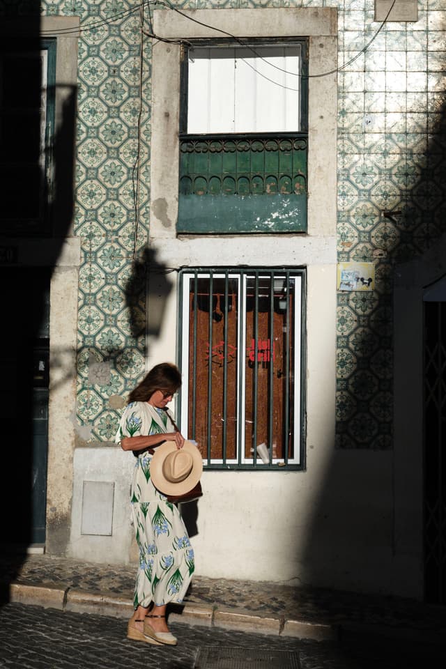 A person in a patterned dress stands in front of a tiled building, holding a hat, with sunlight casting shadows on the wall
