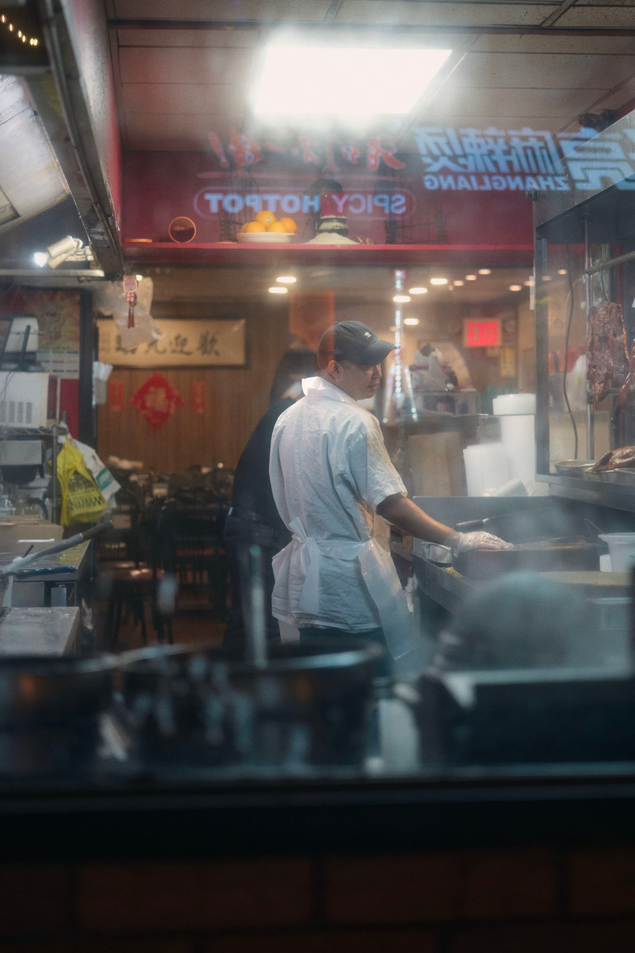 A chef wearing a white apron and black cap cooks in a dimly lit kitchen, with steam rising from the stove. The kitchen has a warm, bustling atmosphere, and there are signs with Chinese characters and Spicy Hotpot visible
