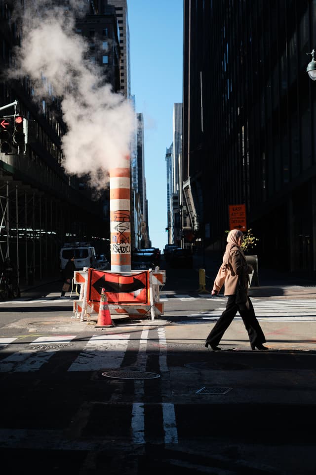 A person walks across a city street with a steam pipe emitting vapor in the background, surrounded by tall buildings and construction barriers