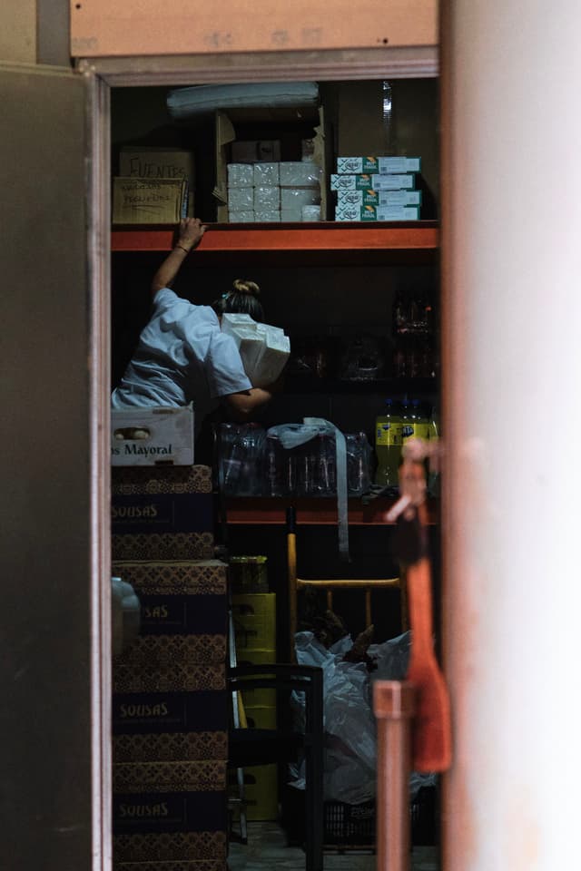 A person reaching for items on a high shelf in a dimly lit storage room, surrounded by boxes and supplies
