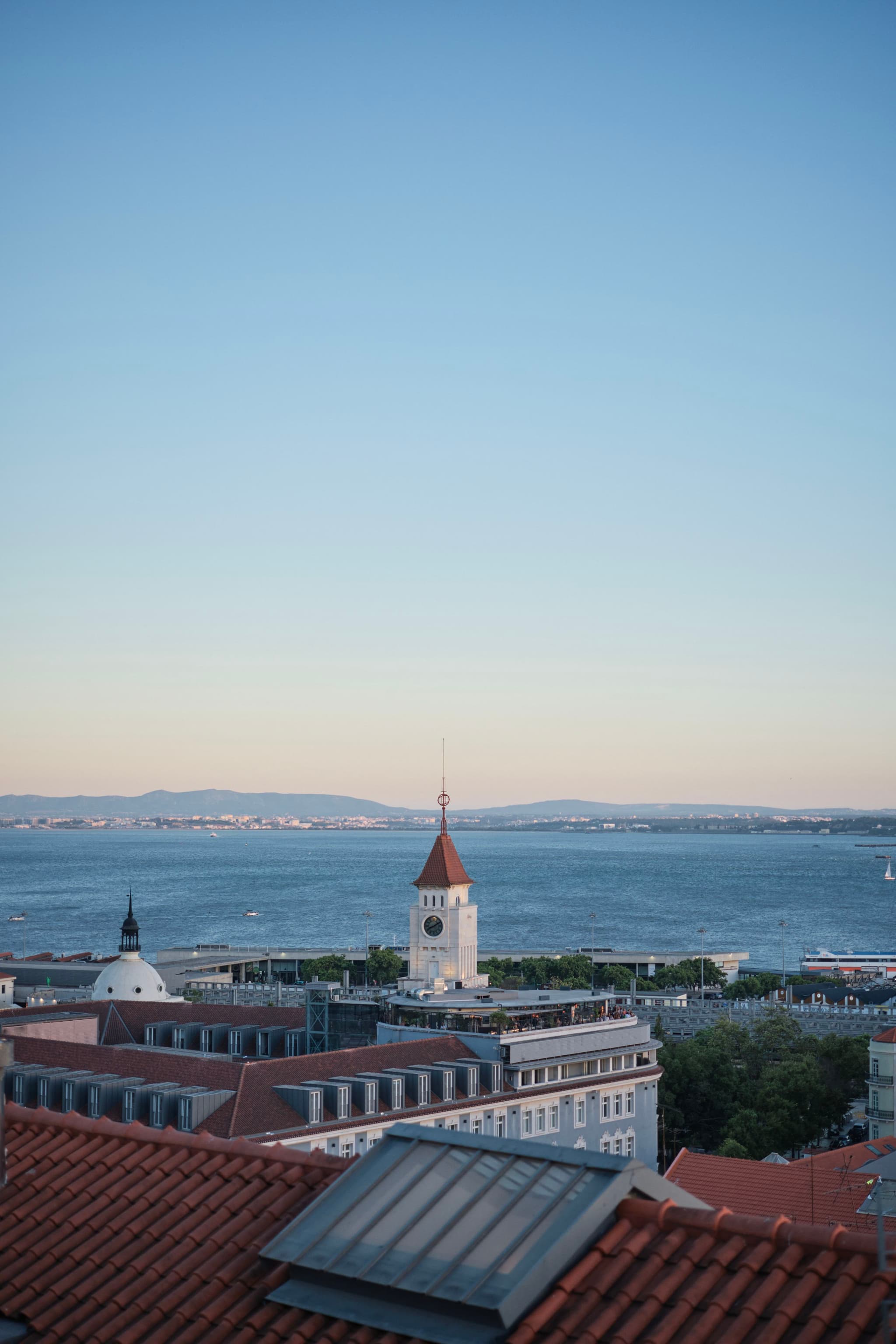 A coastal cityscape with red-tiled rooftops, a clock tower, and a view of the sea under a clear blue sky