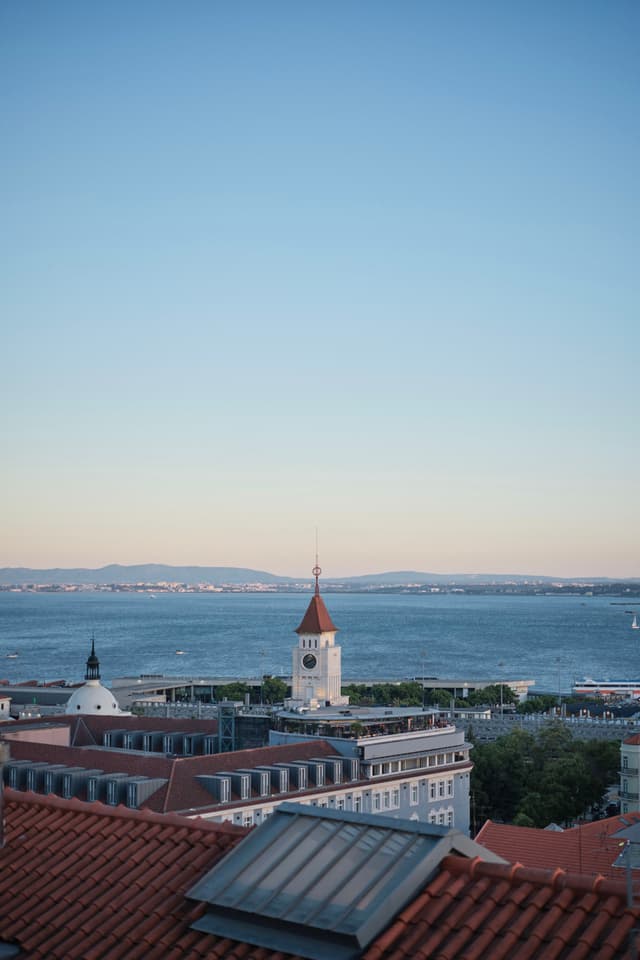 A coastal cityscape with red-tiled rooftops, a clock tower, and a view of the sea under a clear blue sky