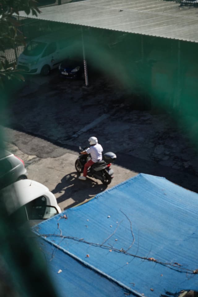 A person wearing a white helmet and shirt is riding a motorcycle in a parking area with several cars and a blue structure nearby