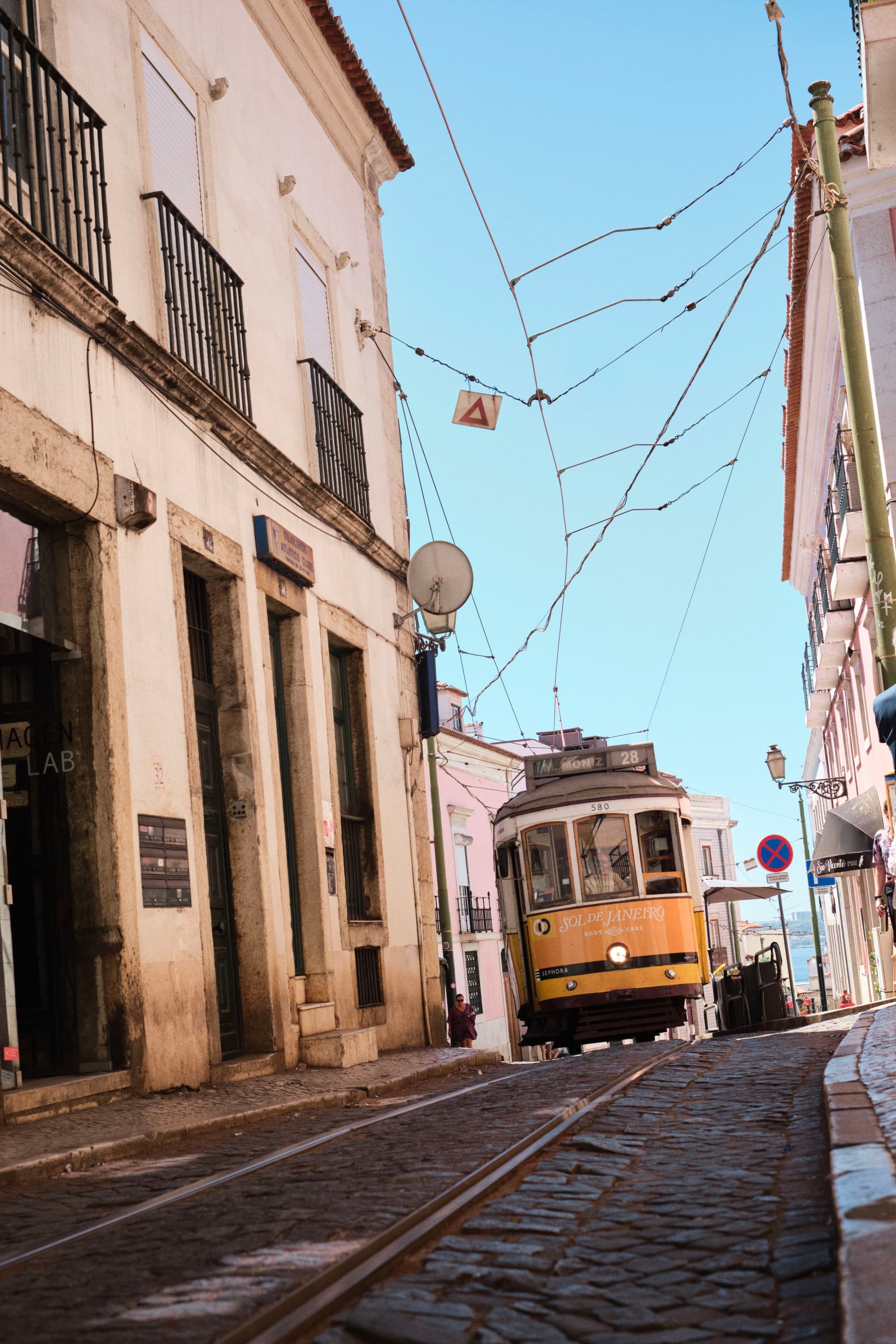 A vintage yellow tram travels up a narrow, cobblestone street lined with old buildings under a clear blue sky