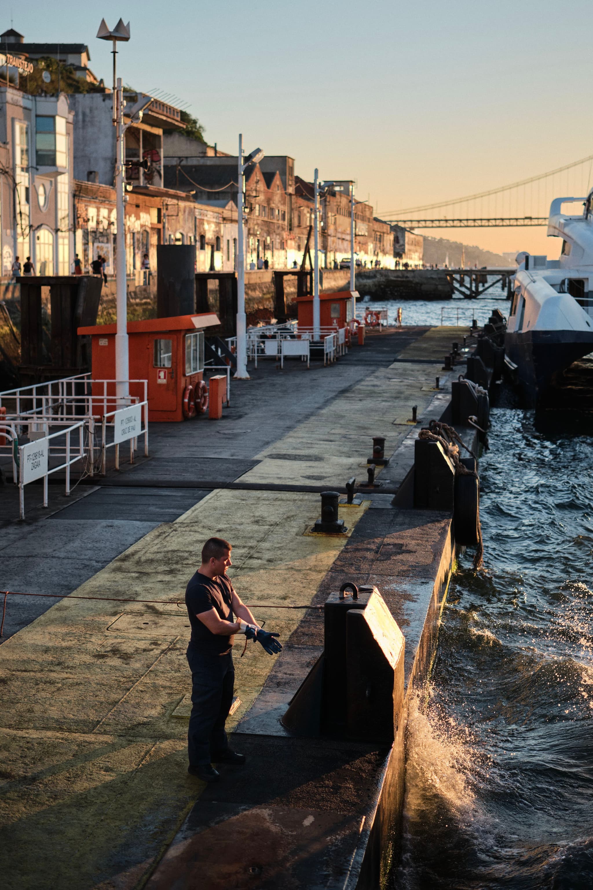 A person stands on a dock near the water, with buildings lining the waterfront and a bridge visible in the background. The scene is lit by the warm glow of the setting sun