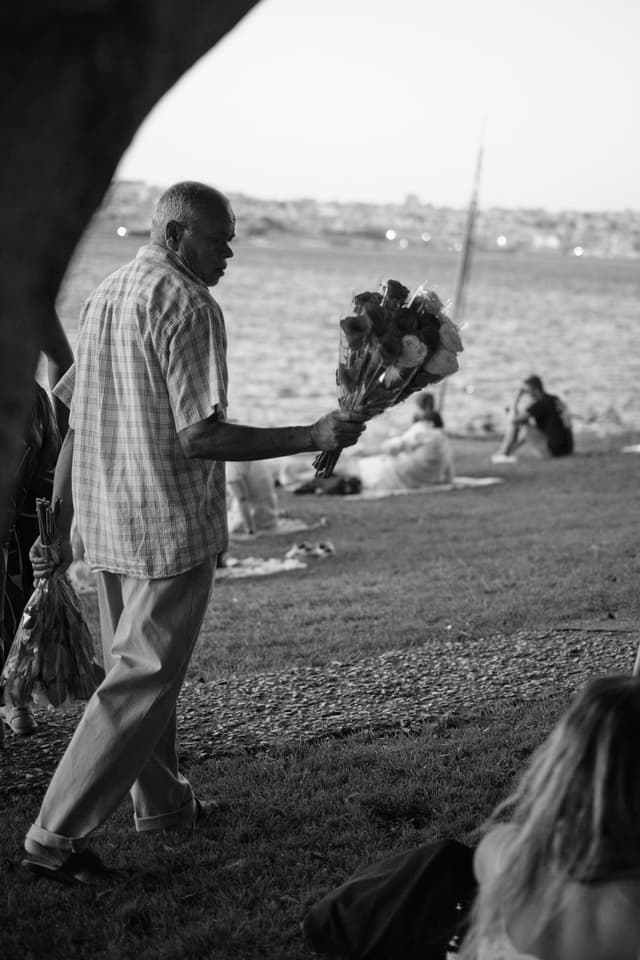 An elderly man holding a bouquet of flowers walks along a grassy area by the water, with people sitting in the background. The scene is in black and white