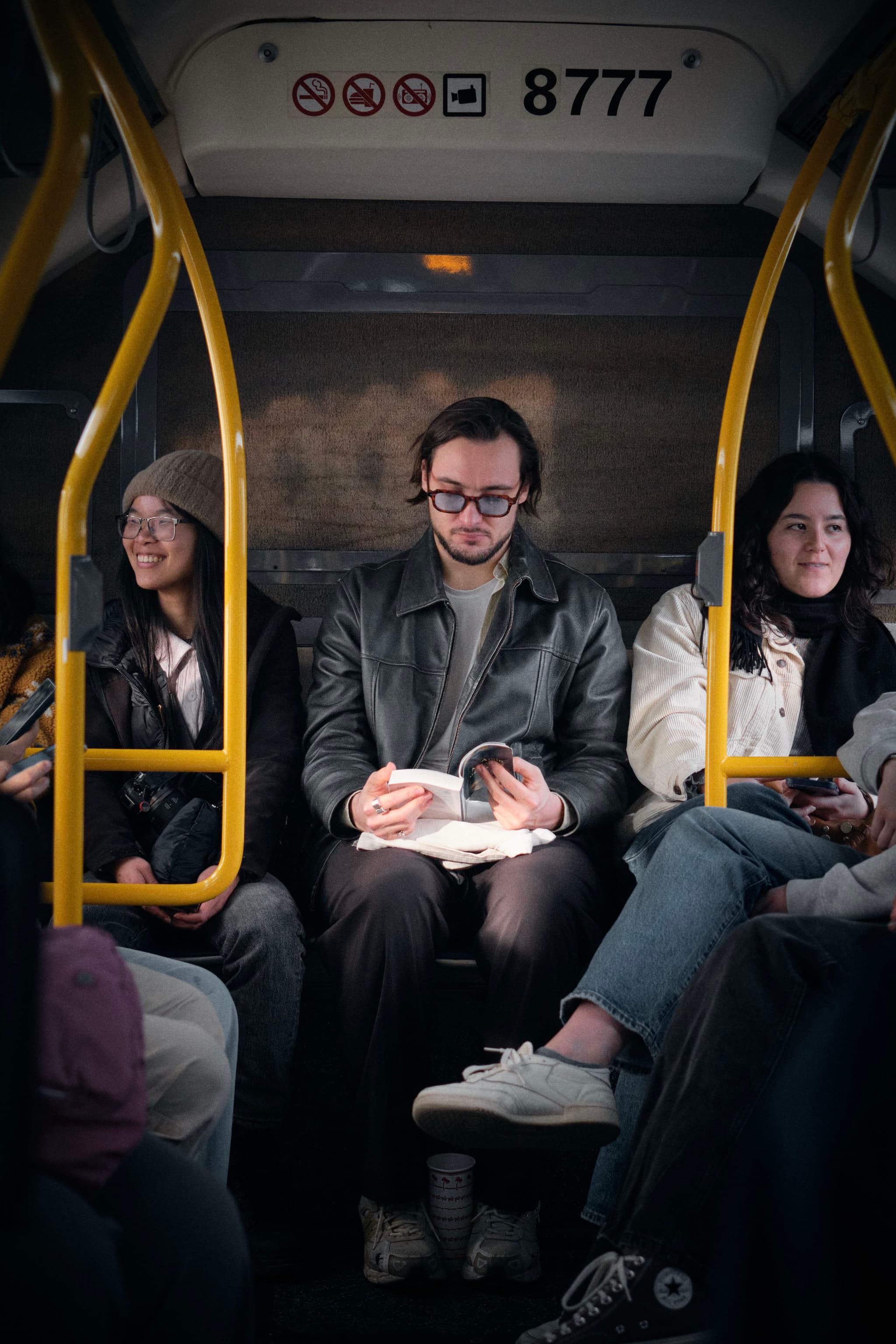 A man reading a book sits between two women on a bus, with yellow handrails and a sign displaying 8777 above them