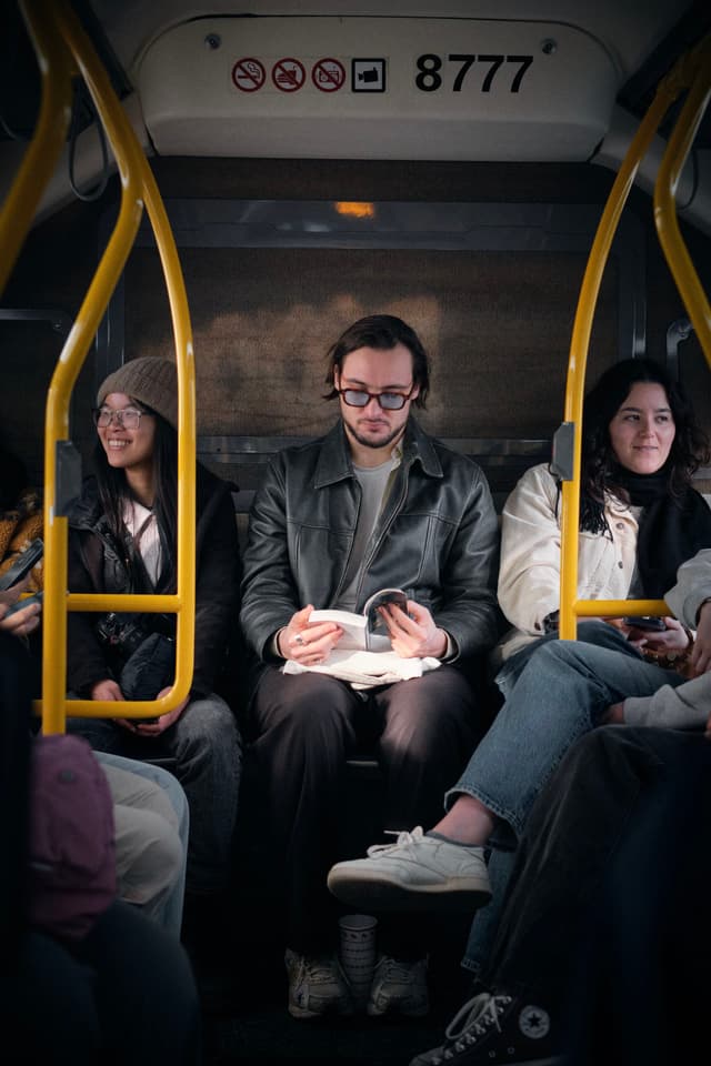 A man reading a book sits between two women on a bus, with yellow handrails and a sign displaying 8777 above them