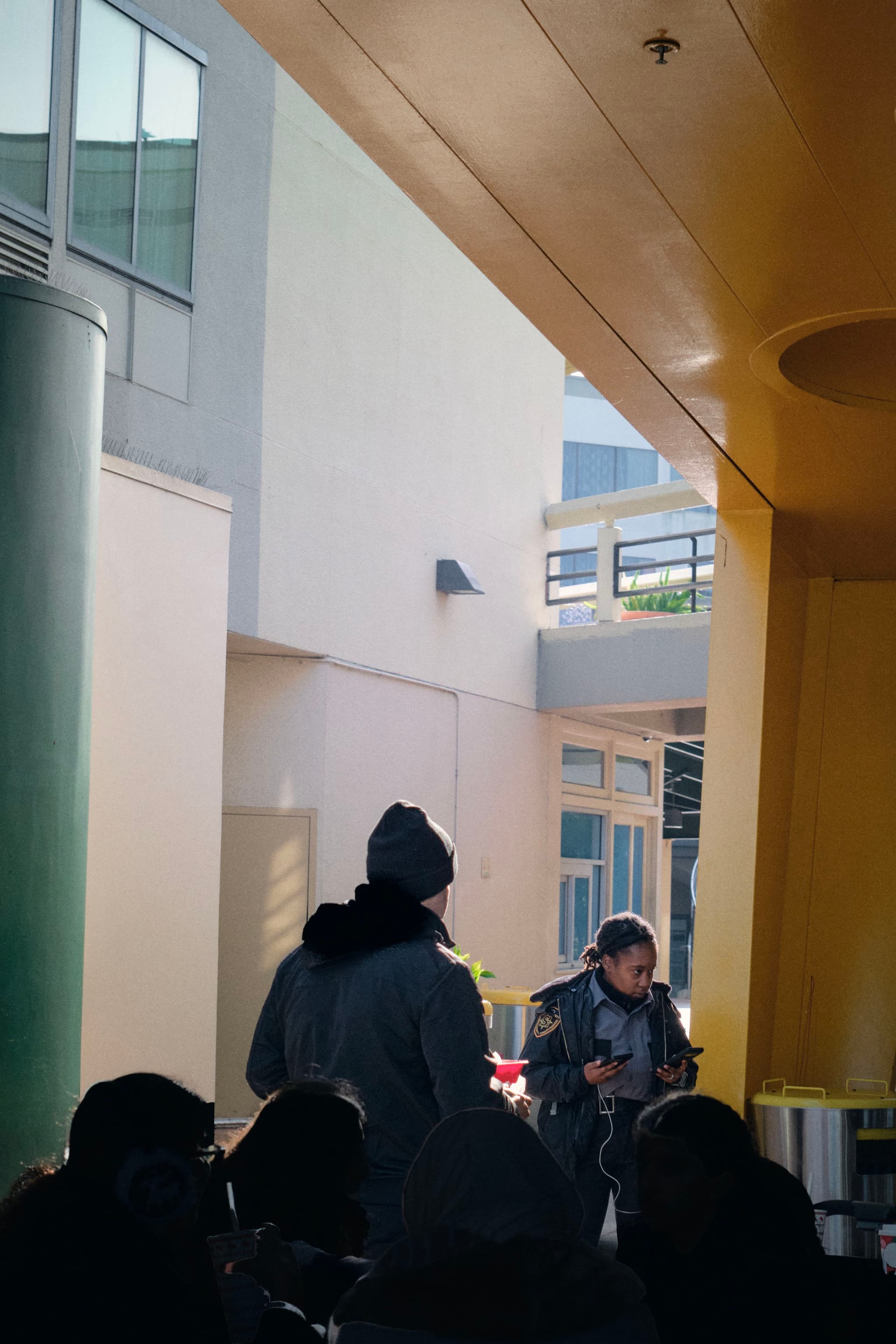 A group of people in an outdoor corridor with a yellow ceiling and green column, some wearing winter clothing
