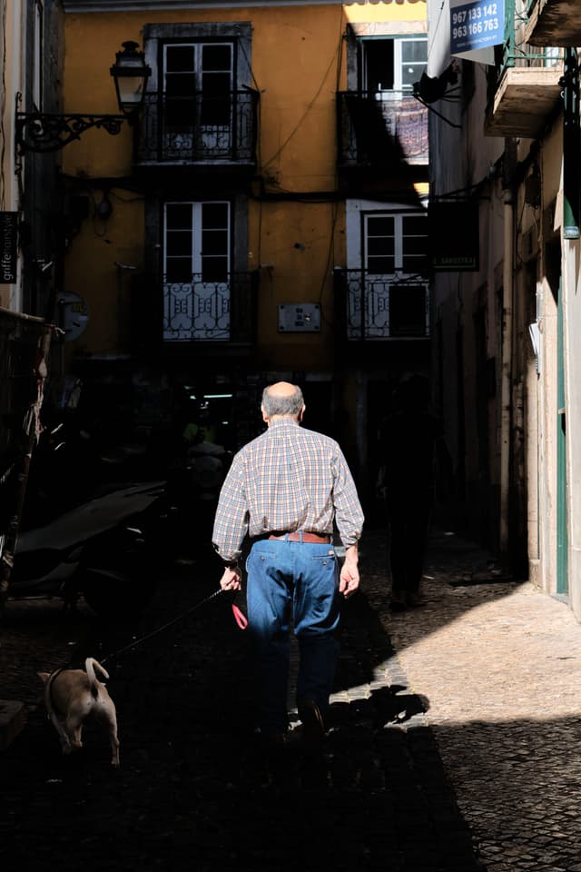 A man walking a dog down a narrow, shadowy street with yellow buildings and balconies in the background