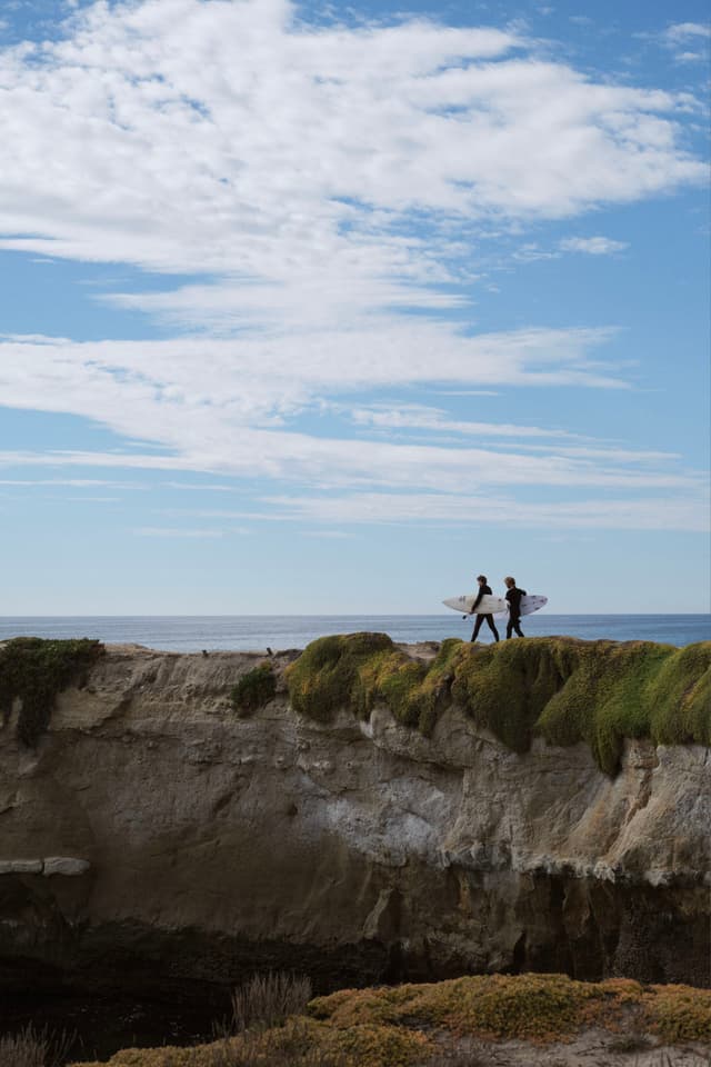 Two people carrying surfboards walk along a grassy cliff edge with the ocean in the background under a partly cloudy sky