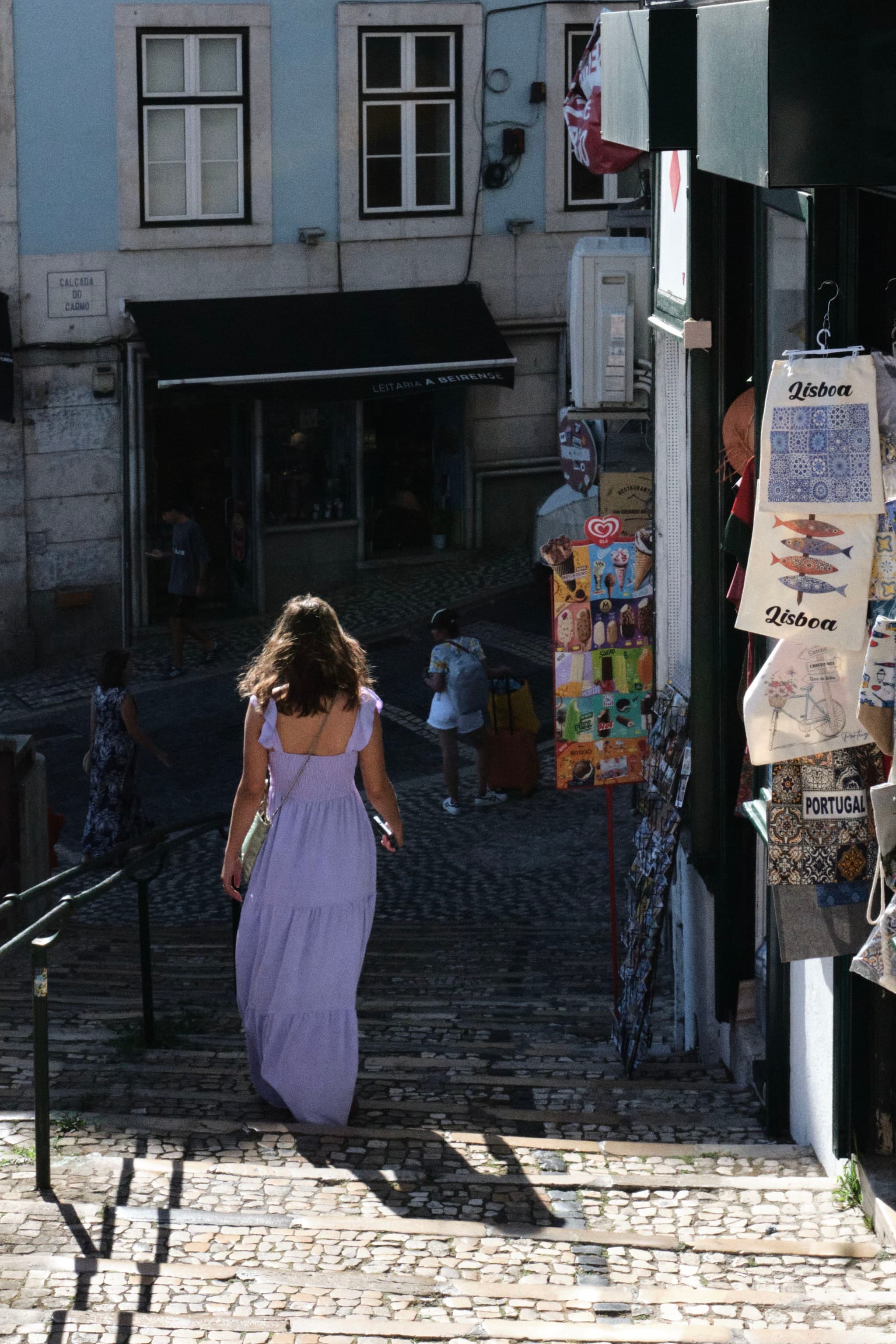 A woman in a long purple dress walks down a cobblestone street lined with shops and colorful posters, with sunlight casting shadows on the ground