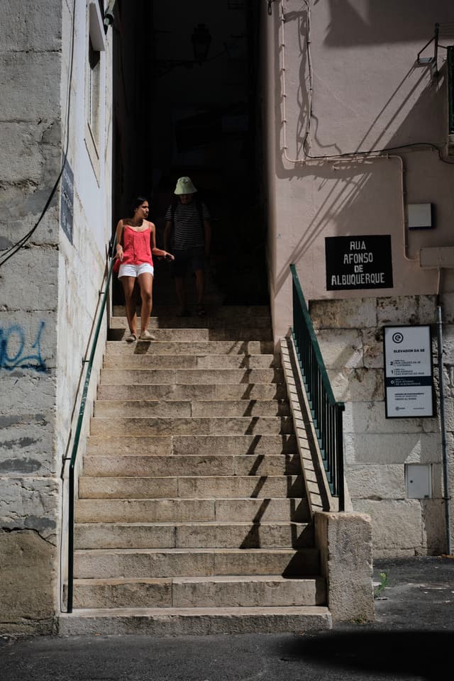 A woman in a pink top and white shorts descends a narrow stone staircase between buildings, with a man in a hat following behind. The scene is partially shaded, and there are signs on the wall