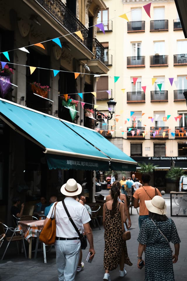 A lively street scene with people walking under colorful bunting, surrounded by buildings with balconies