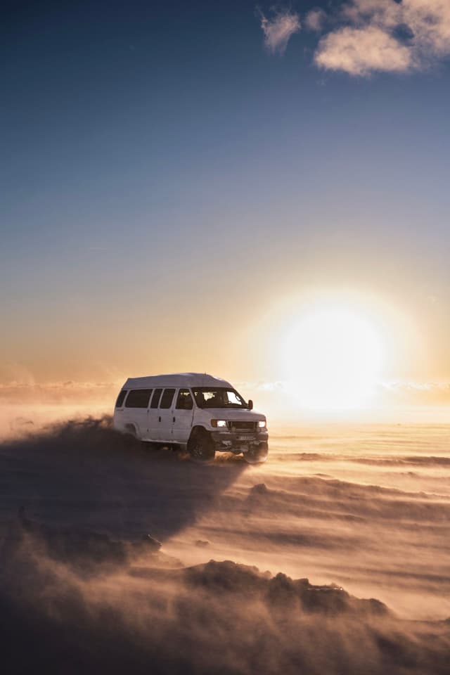A white van drives through a snowy landscape at sunset, with the sun low on the horizon and clouds of snow swirling around
