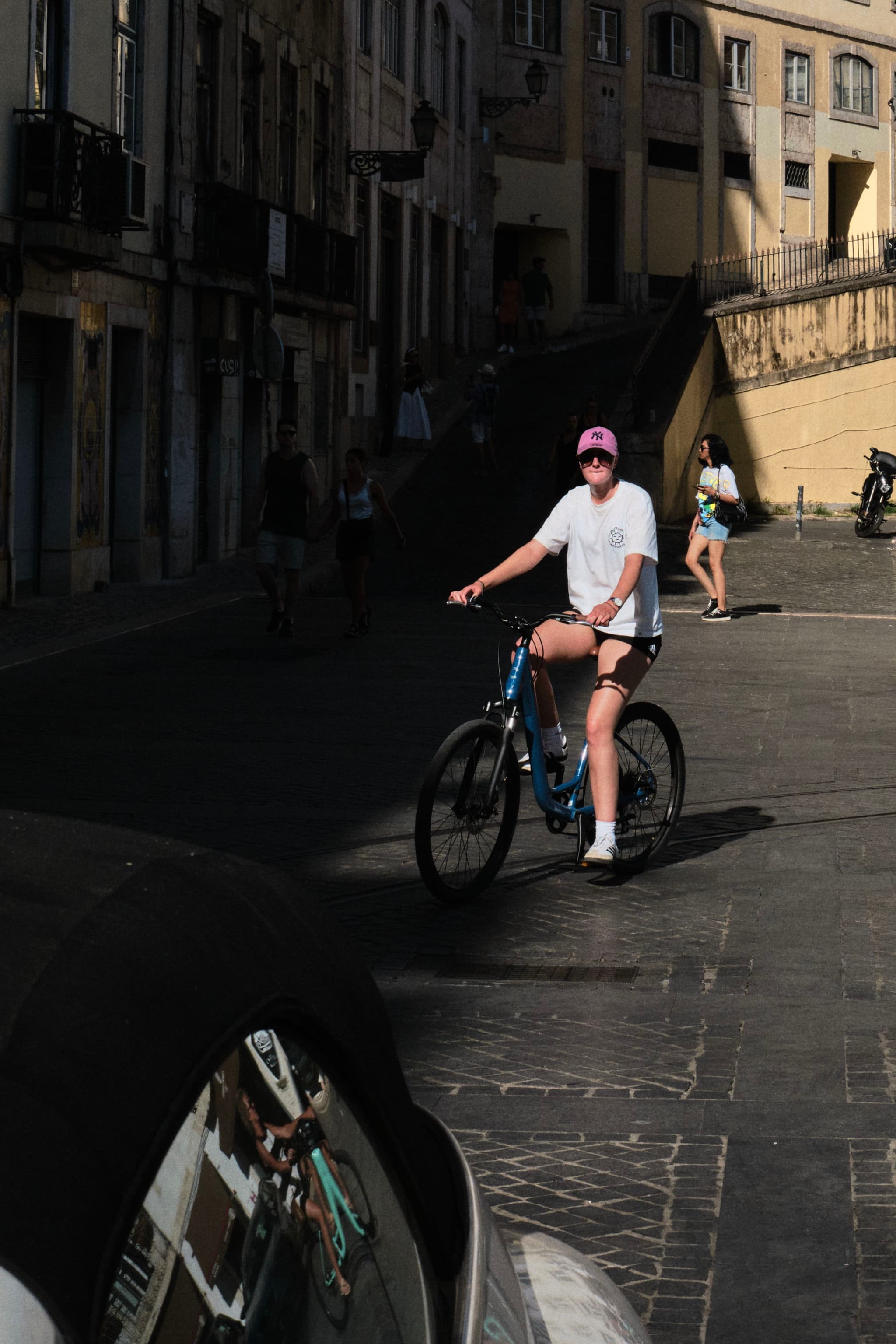 A person rides a bicycle on a sunlit street, with buildings and another person in the background