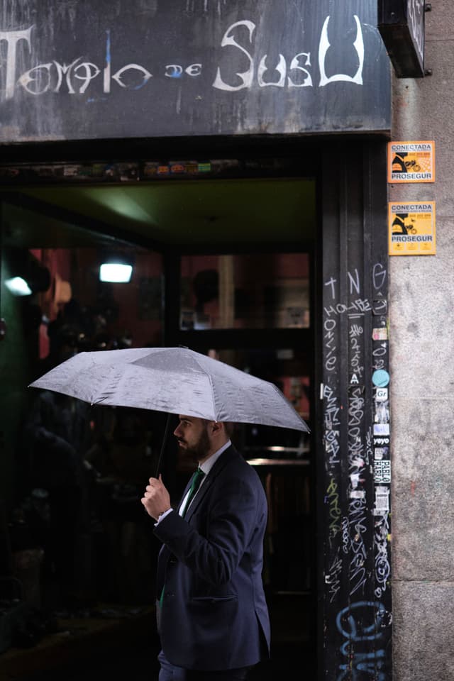 A person in a suit holds an umbrella while standing outside a graffiti-covered entrance