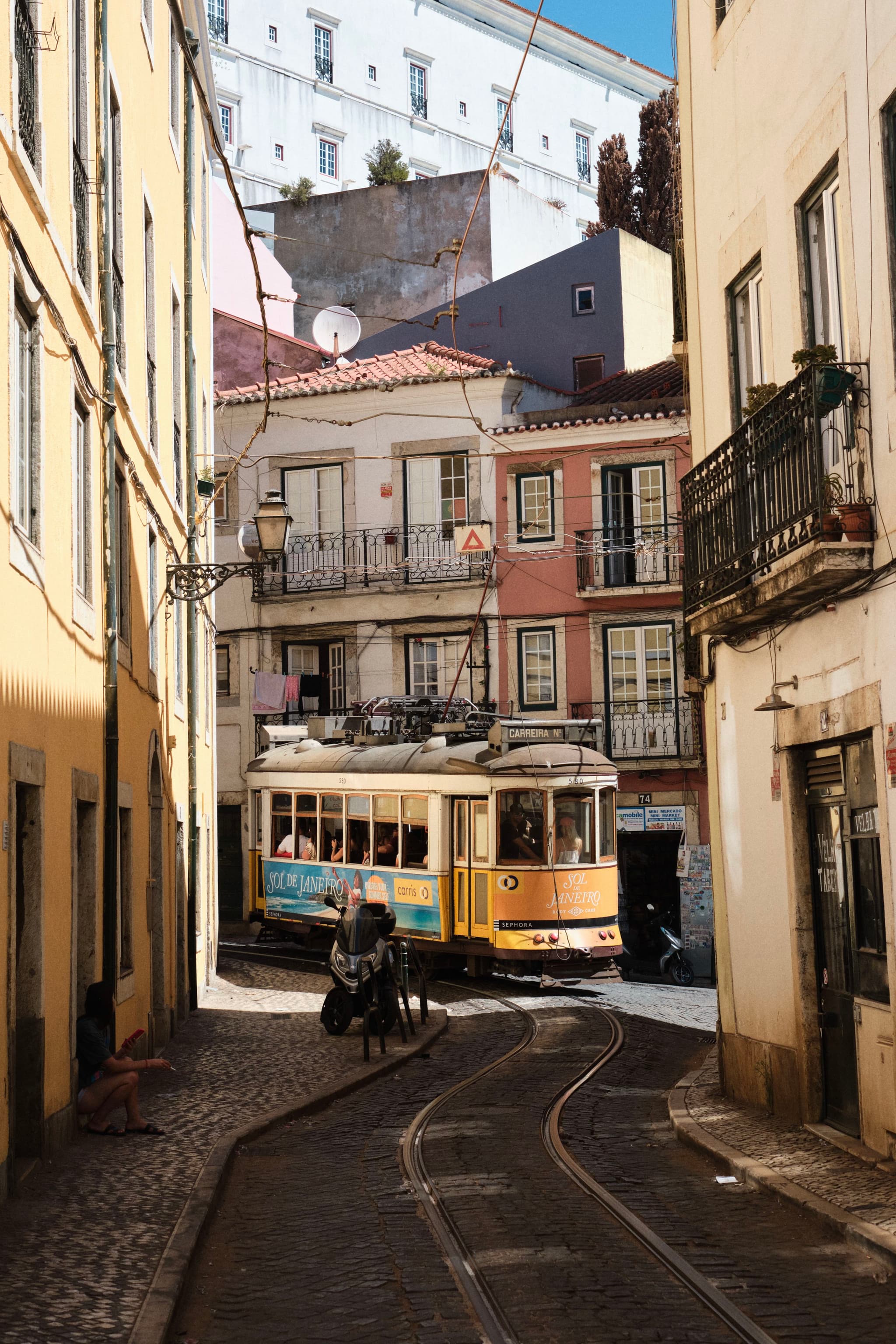 A yellow tram navigates a narrow, cobblestone street lined with colorful buildings