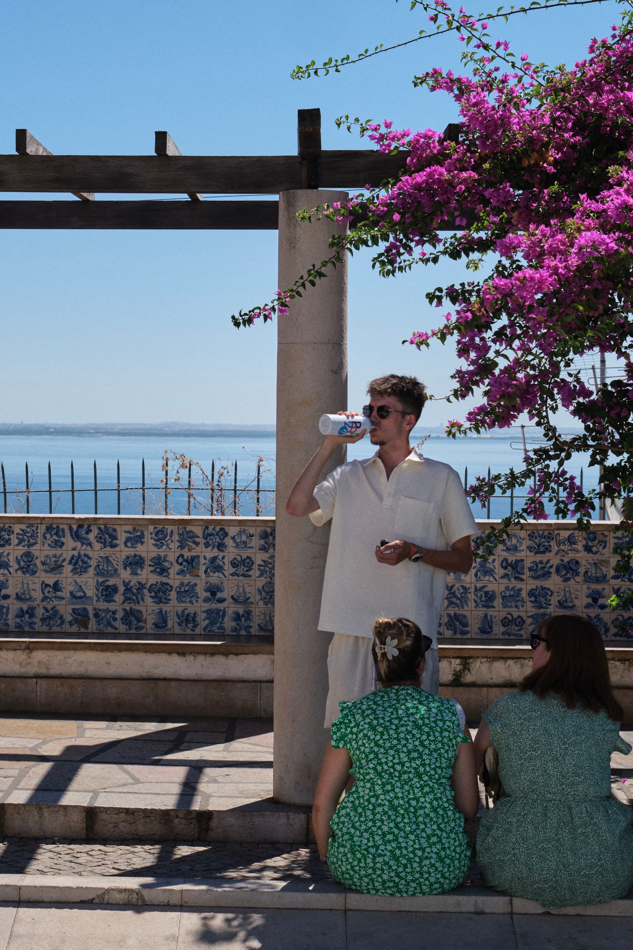 A person in light clothing drinks from a bottle while standing near a pergola with vibrant pink flowers. Two people in green dresses sit on the ground, with a scenic view of the ocean in the background