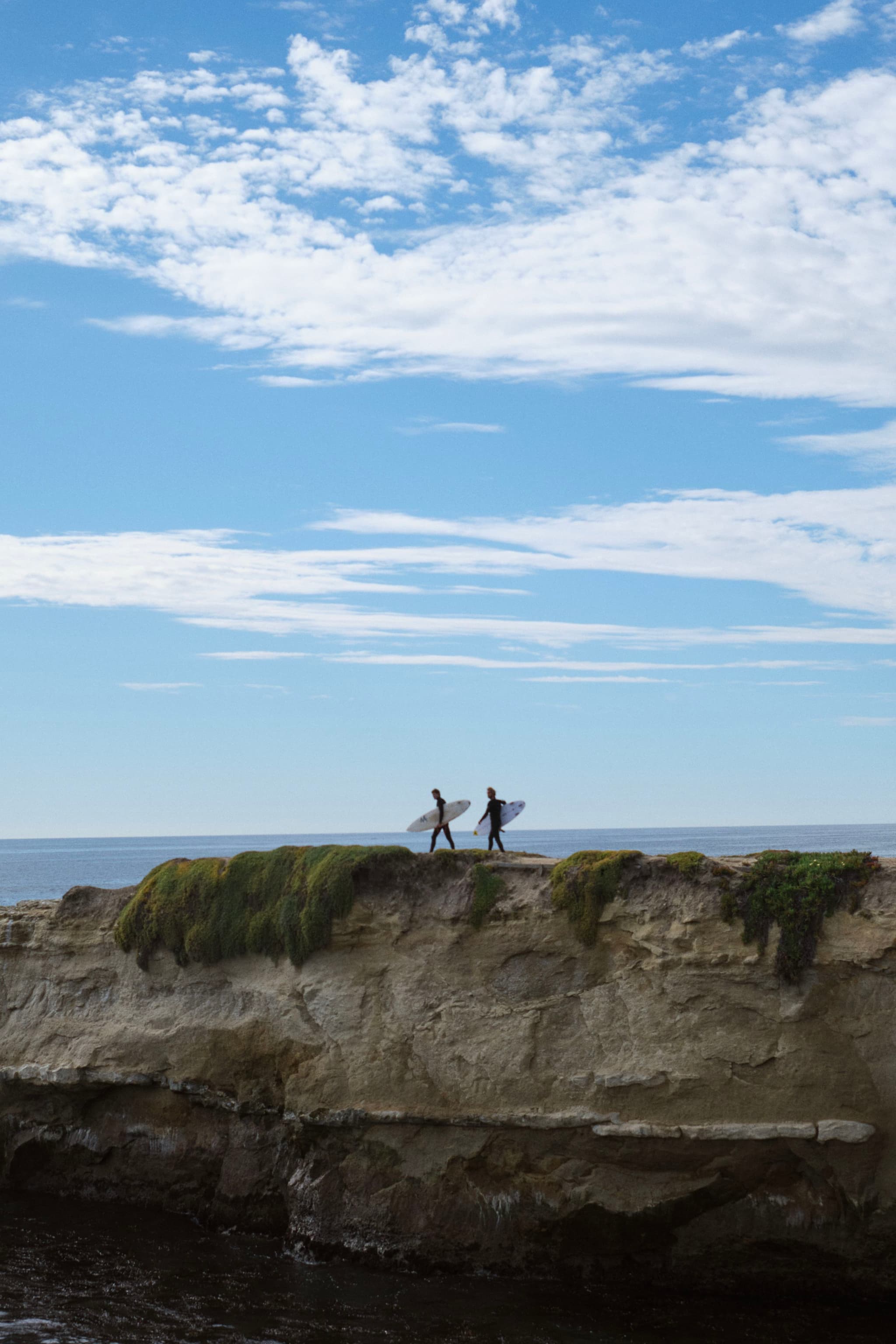 Two people carrying surfboards walk along a rocky cliff with the ocean and a partly cloudy sky in the background