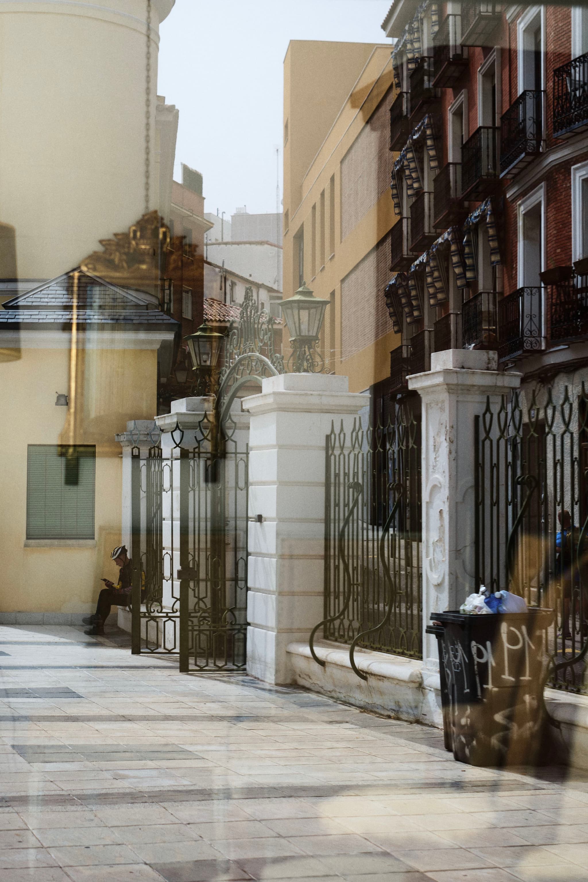 A street scene with a mix of architectural styles, featuring wrought iron gates, brick buildings, and a person sitting in the background. The image has a reflective quality, possibly due to a glass surface