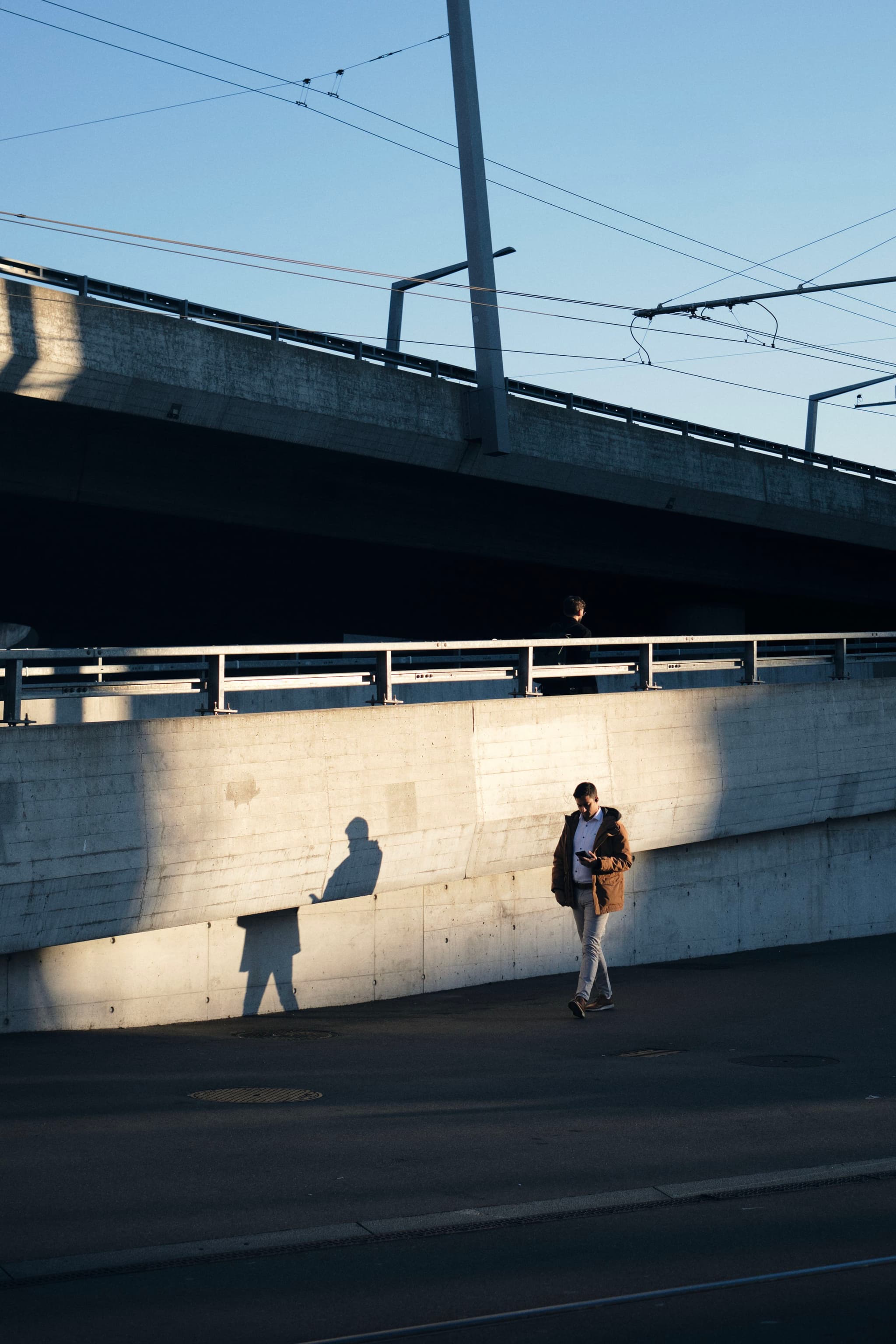 A person walking along a sunlit concrete wall with a shadow cast beside them, under an overpass with visible power lines