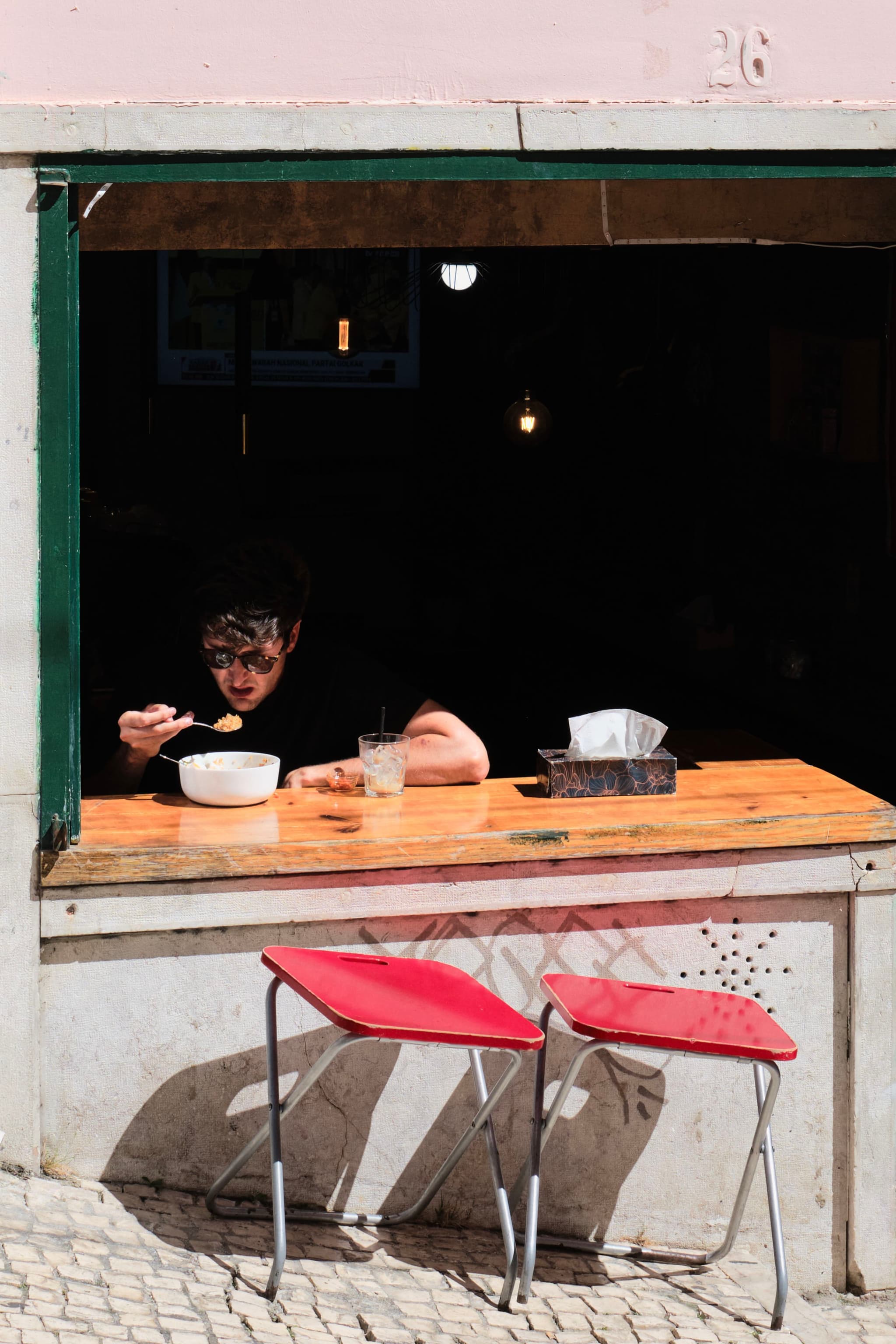 A person eating at a wooden counter inside a window opening, with two red chairs outside on a cobblestone street