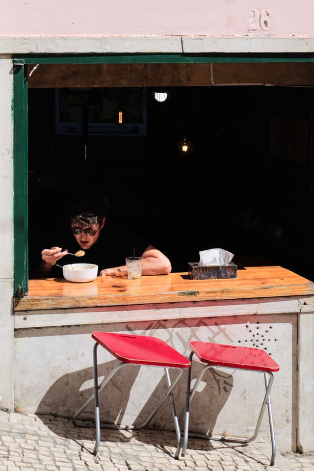 A person eating at a wooden counter inside a window opening, with two red chairs outside on a cobblestone street