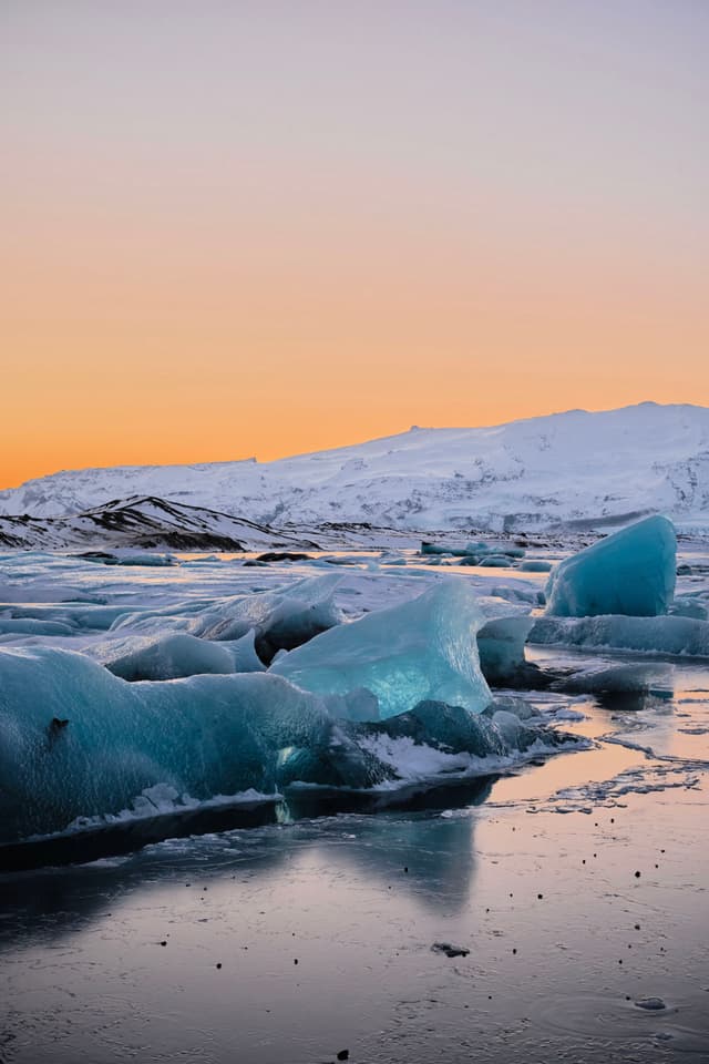 A serene icy landscape with large blue ice formations on a reflective water surface, set against a backdrop of snow-covered mountains and a warm, orange sunset sky