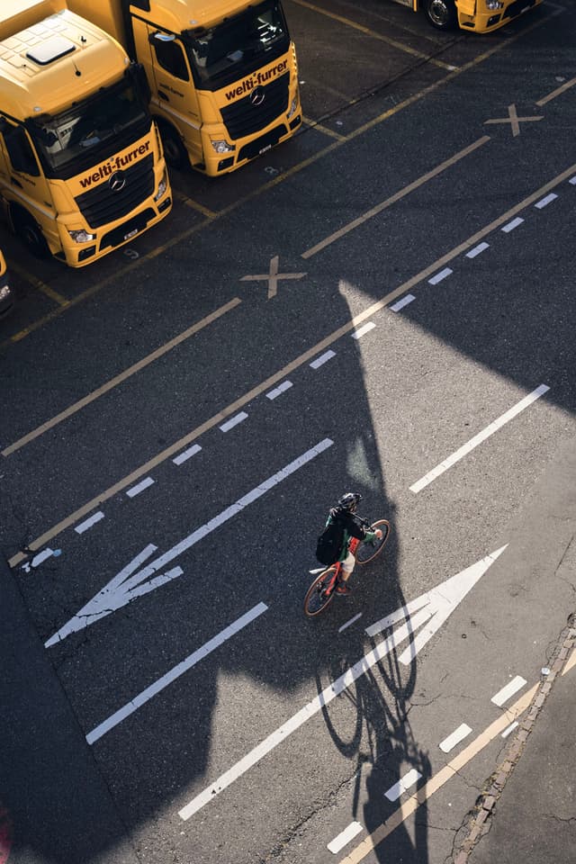 A cyclist rides along a road with marked lanes, casting a shadow. Yellow trucks are parked in a lot nearby