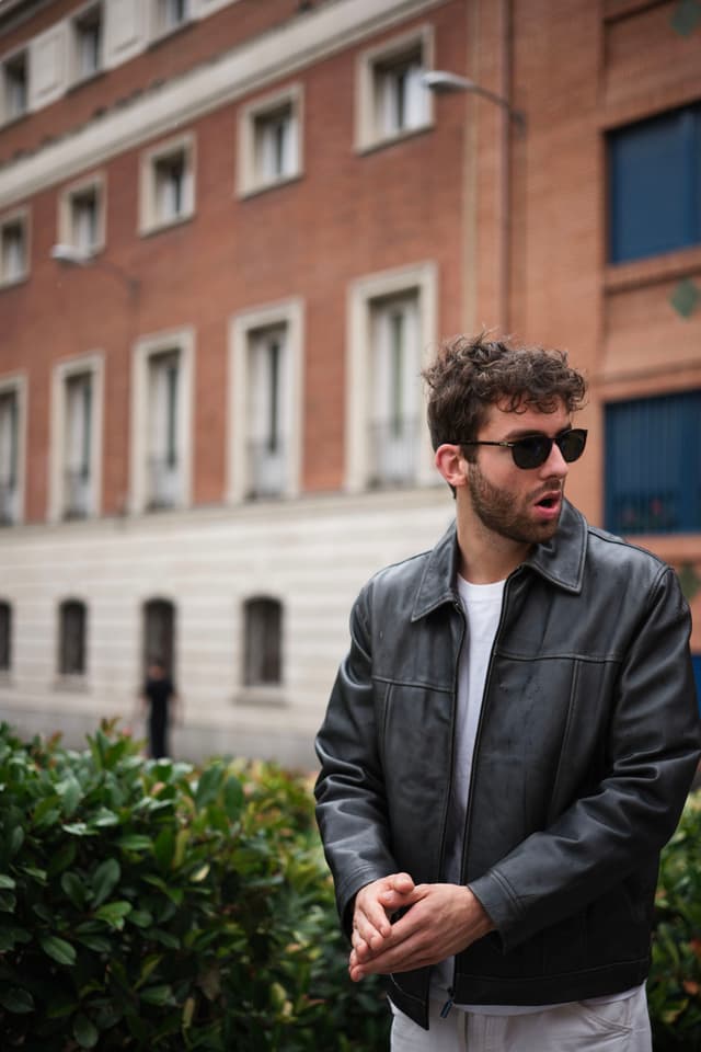 A man wearing sunglasses and a leather jacket stands in front of a brick building, with greenery in the foreground