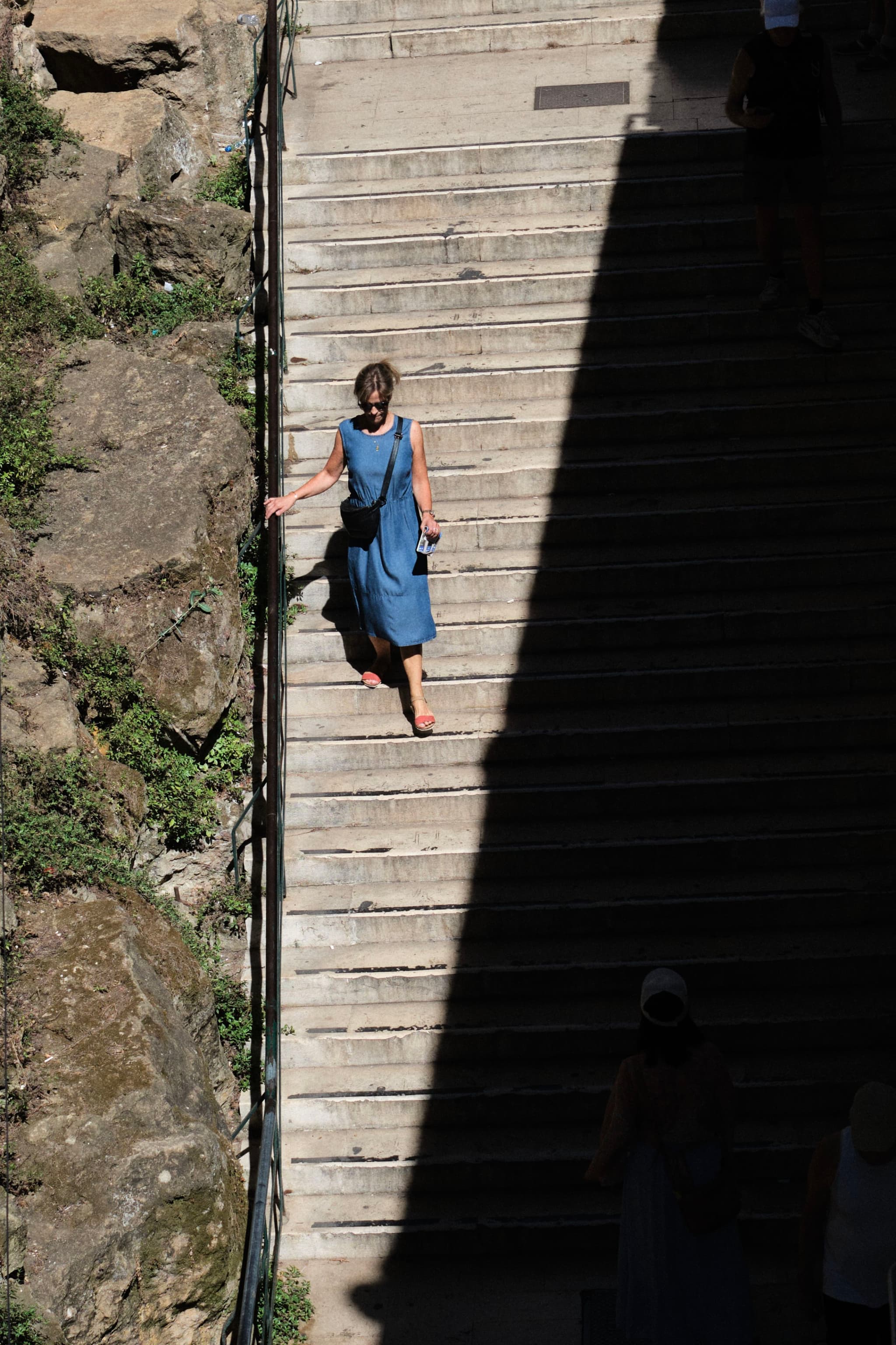 A person in a blue dress walks down a set of stone stairs, partially in shadow, with rocky terrain and greenery on the side