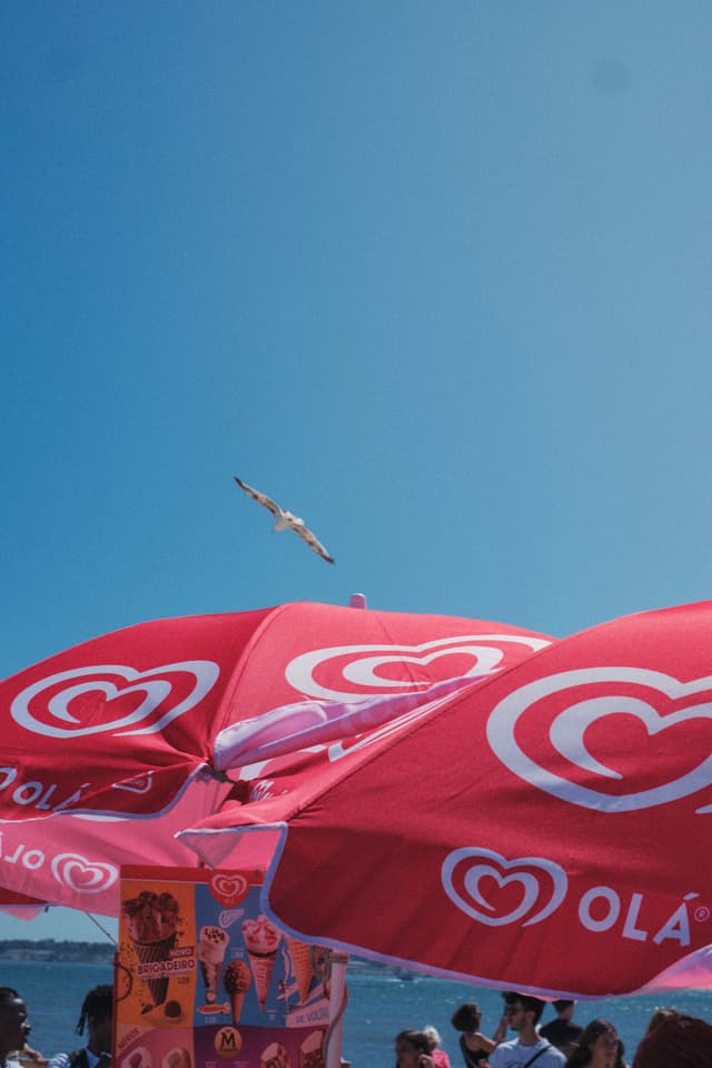 Red beach umbrellas with a heart logo, a seagull flying above, and a clear blue sky